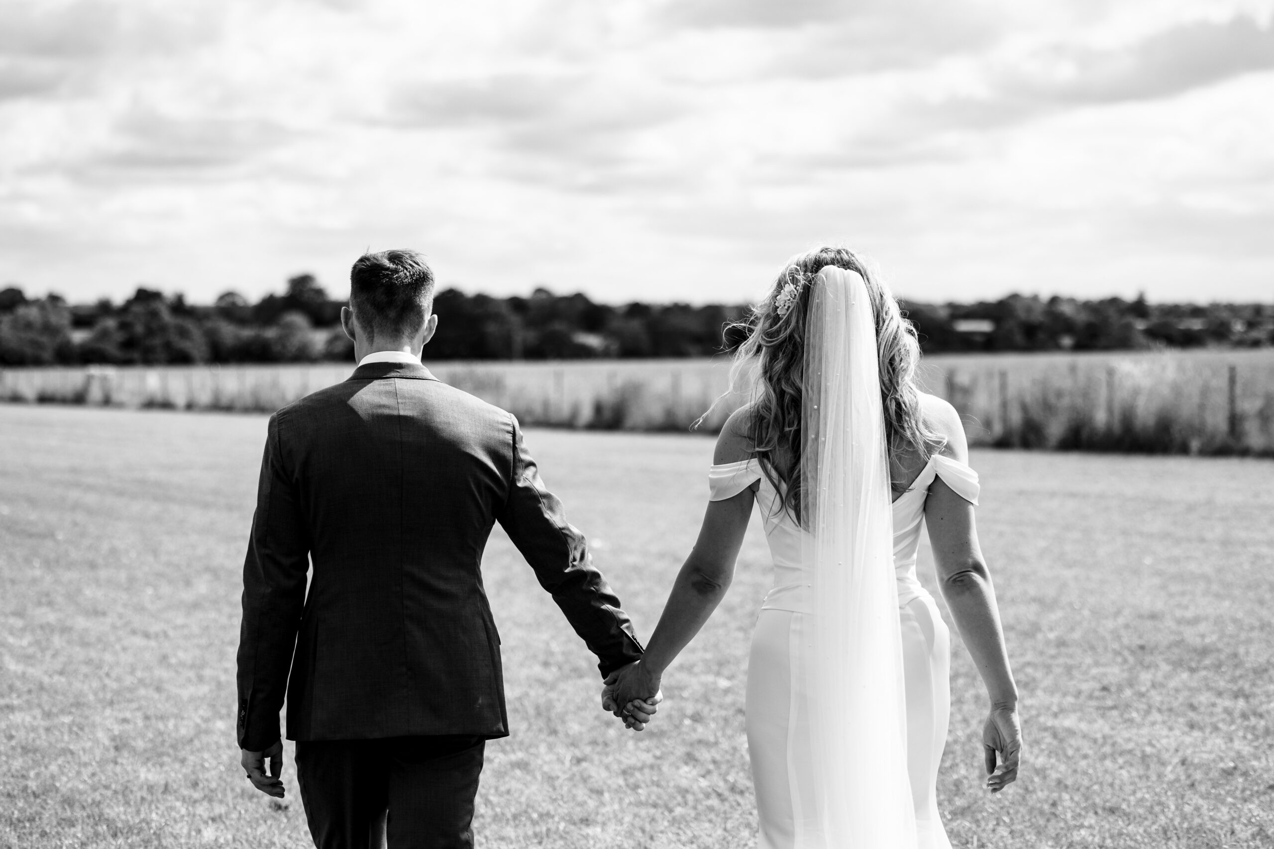 A black and white image of the back of the bride and groom. They are holding hands and there is a field in the distance.