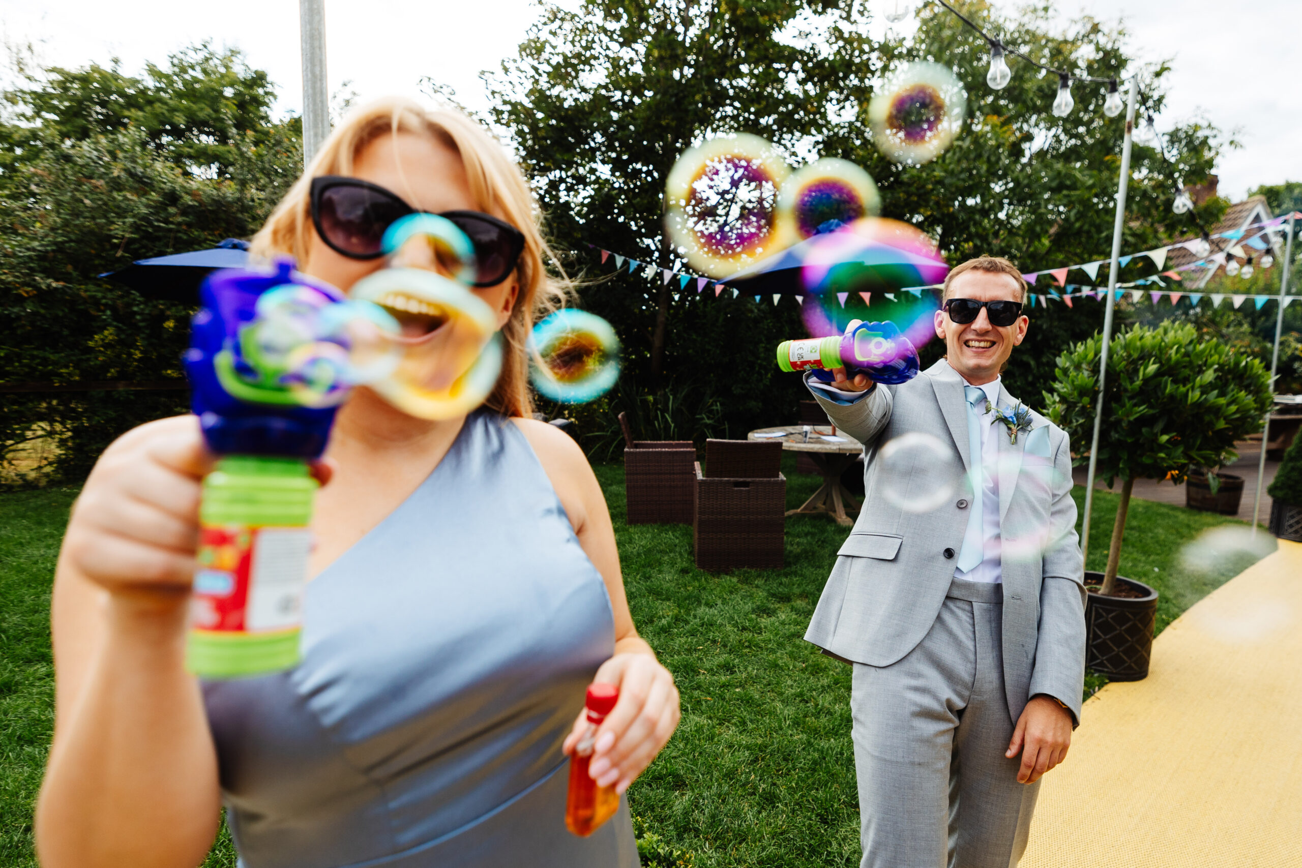 A bridesmaid and member of the groom's party with bubble machines. The bubbles are in the air and the two are smiling.