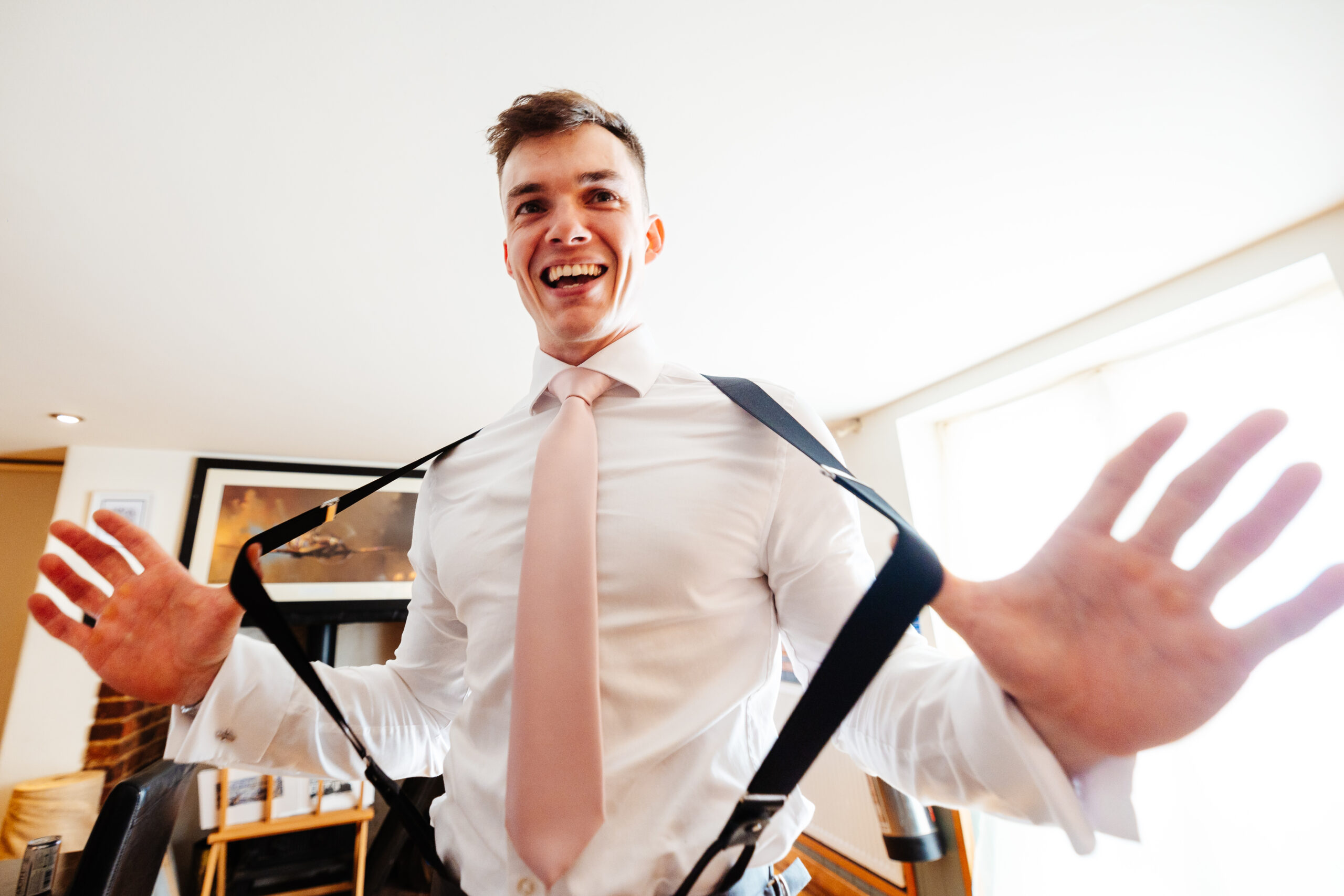 James, the groom in a white shirt and pink tie pulling his braces apart. He has a smile on his face.