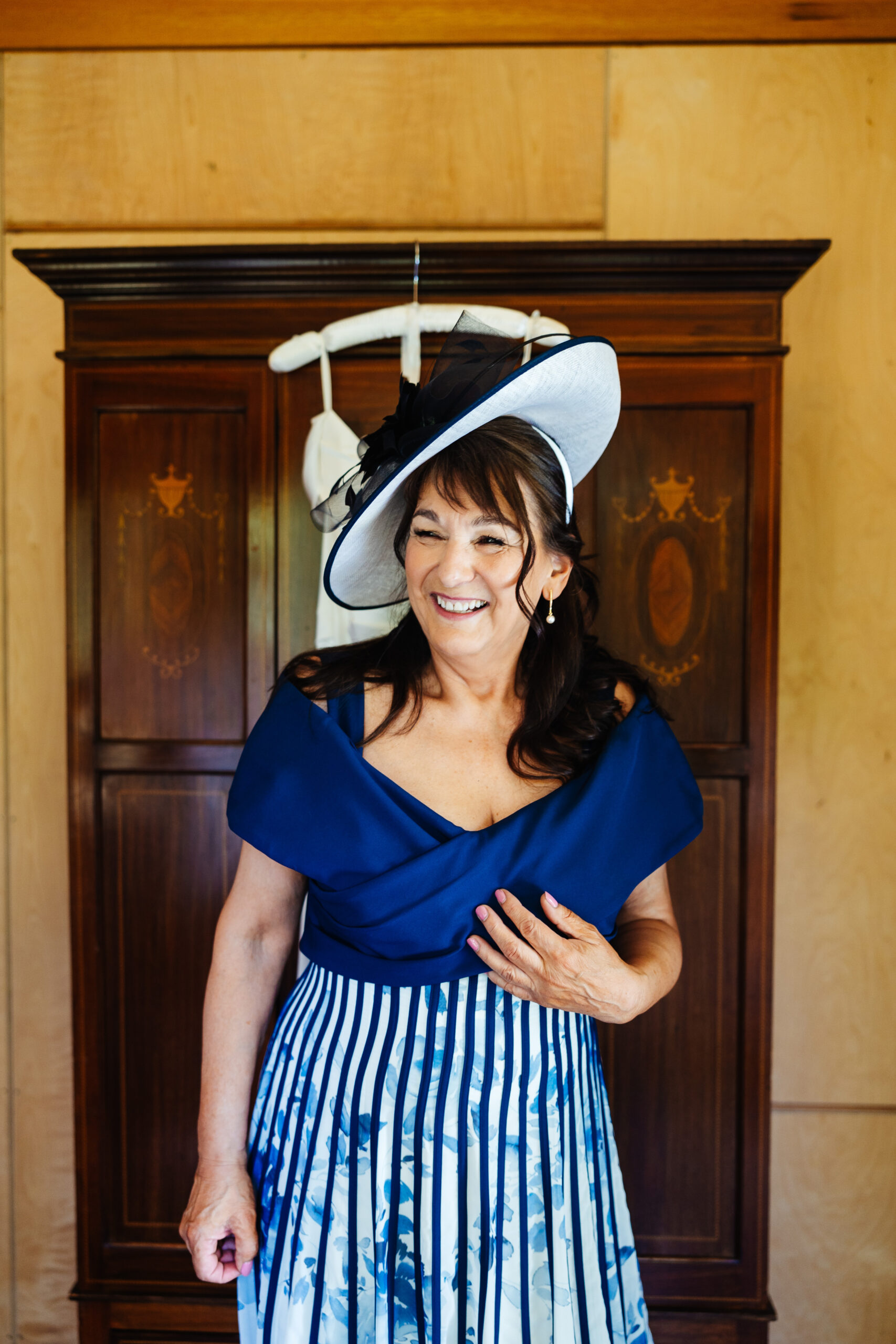 The mother of the bride in a navy blue dress. The bottom of the dress is pleated. She is wearing a matching hat. She is looking away from the camera and she is smiling.