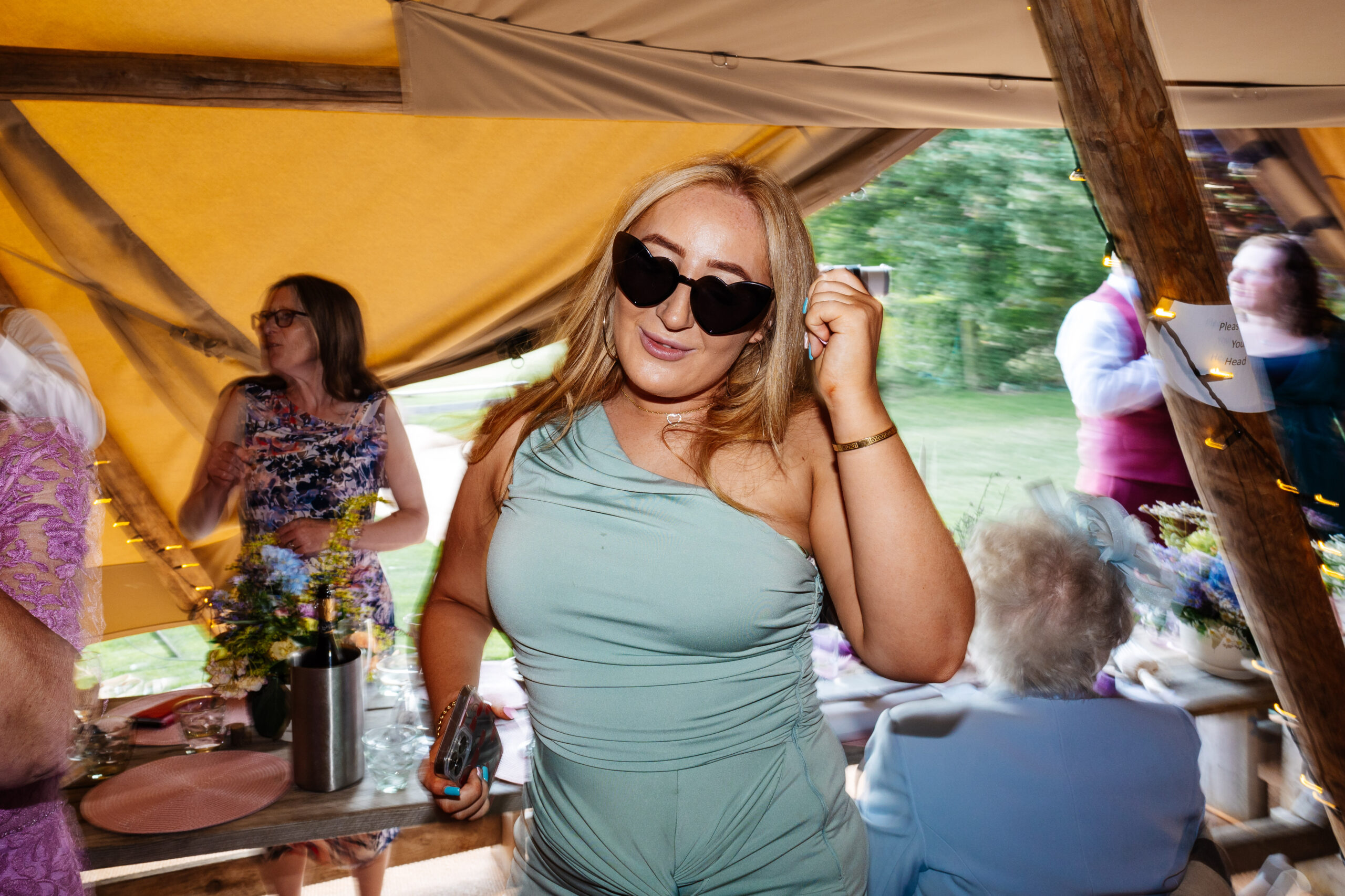 A guest in a turquoise dress dancing - she has black heart sunglasses on.