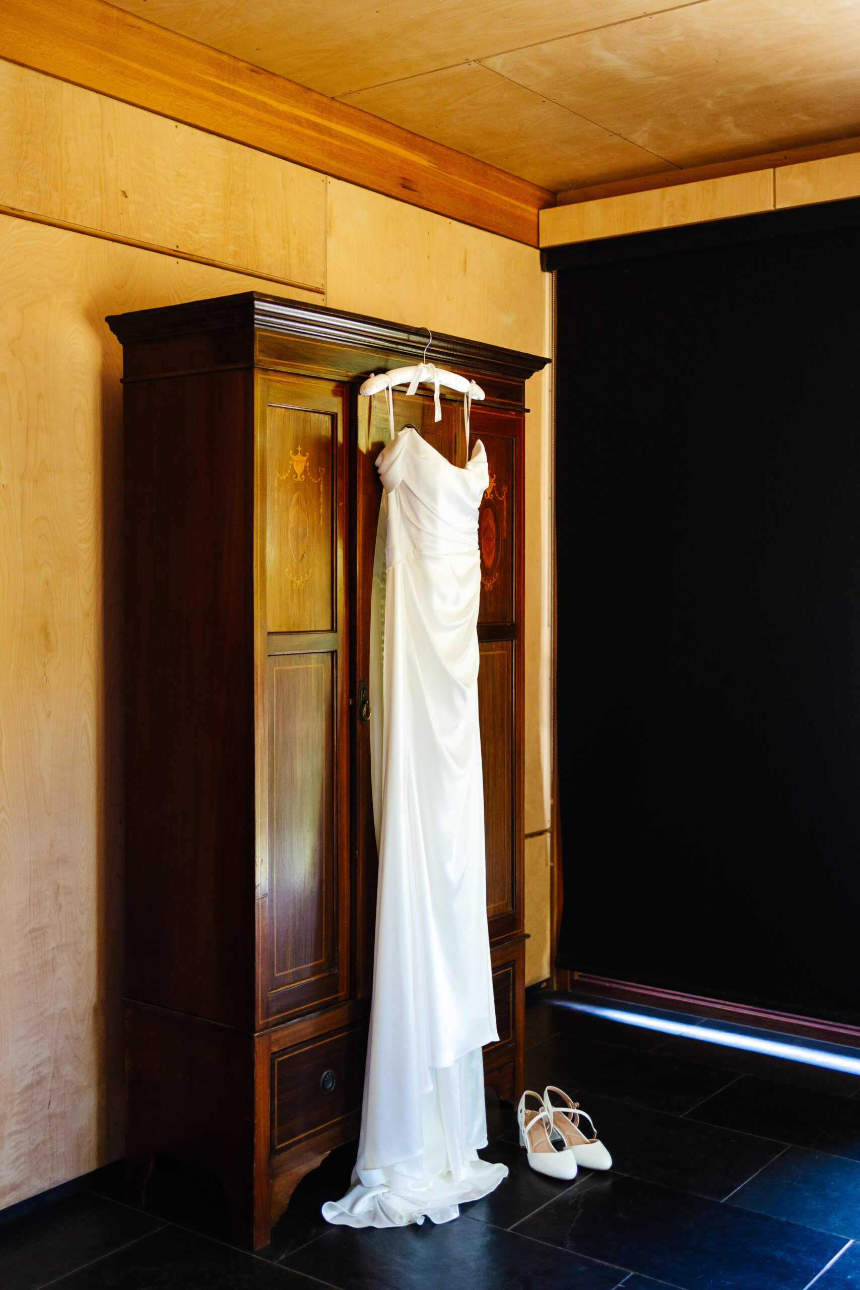 A photo of the bride's wedding dress hung up on an old, wooden wardrobe. It is hanging on a white hanger with white ribbon hanging down. Her shoes are next to the dress.