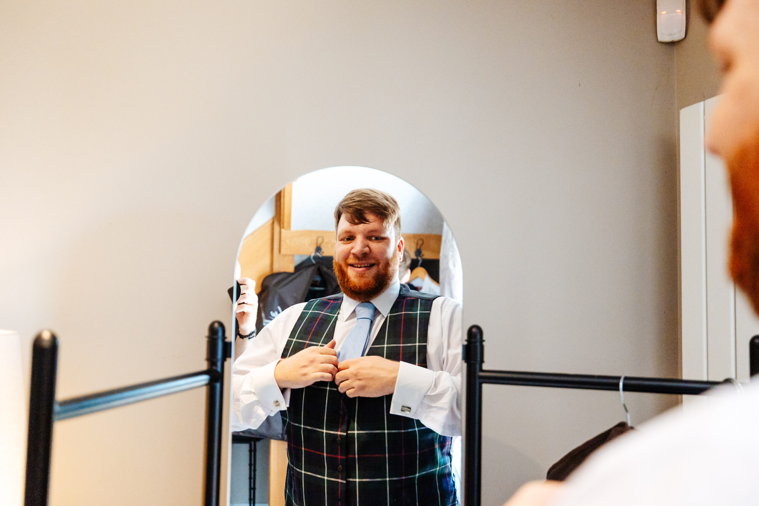 The groom looking at himself in the mirror. He is smiling and is wearing a Scottish print waistcoat.