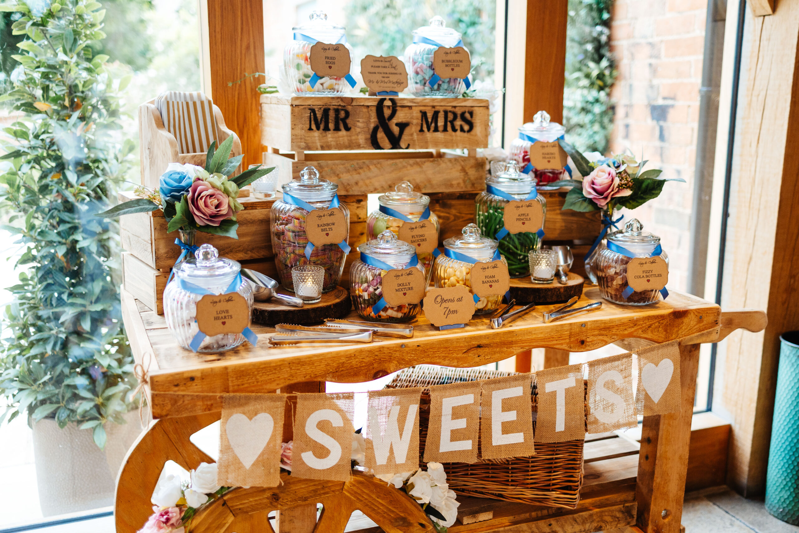 A rustic looking table containing lots of sweet jars with blue ribbon around them telling the guests what is inside them. There are old school, striped paper bags on the table and a sign saying Mr & Mrs.