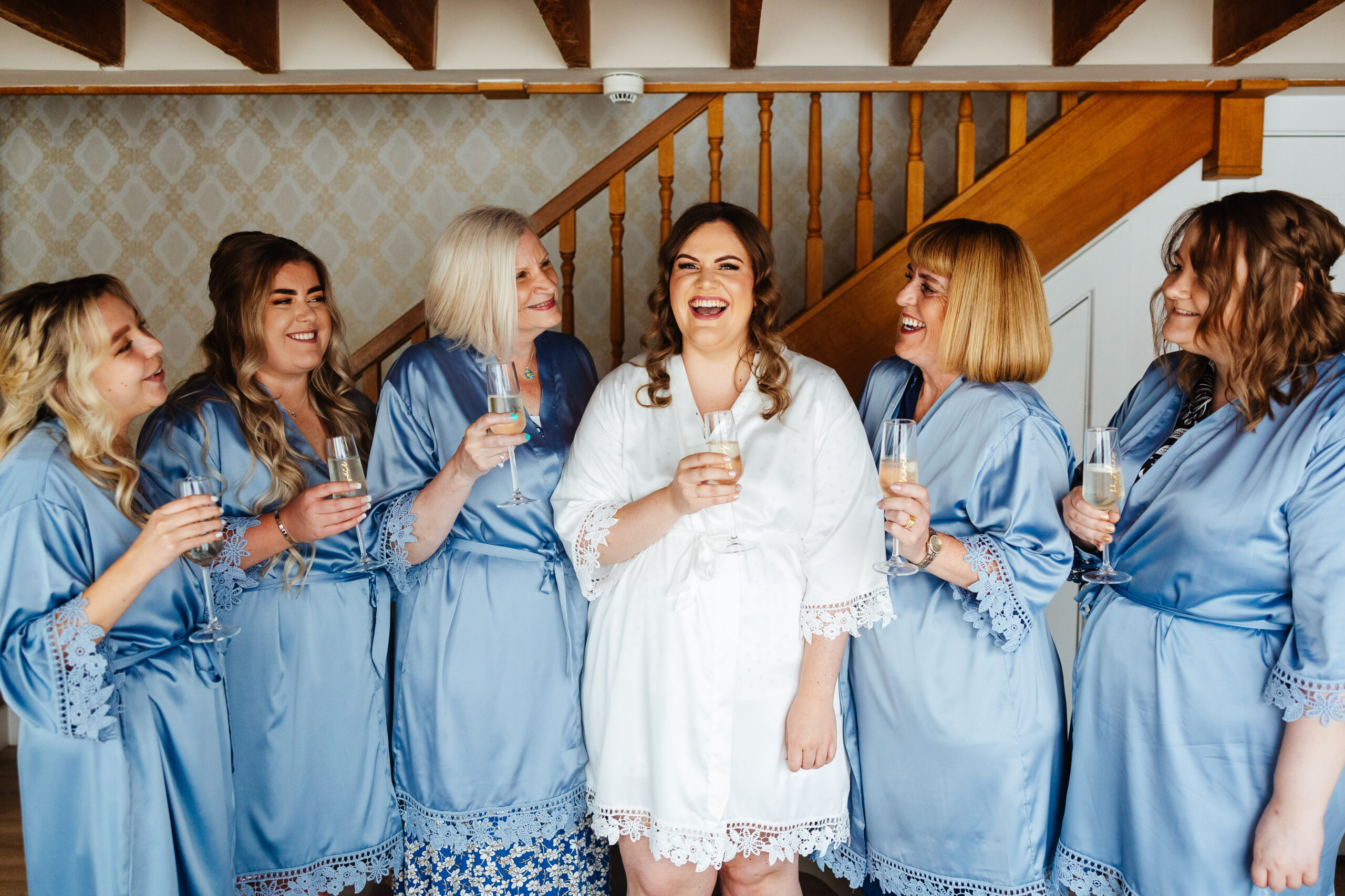 The bride and her bridesmaids. They are stood in the hallway and are wearing blue silk dressing gowns, apart from the bride who is in a white one. They are holding Prosecco and smiling. The bridesmaids are looking at the bride.