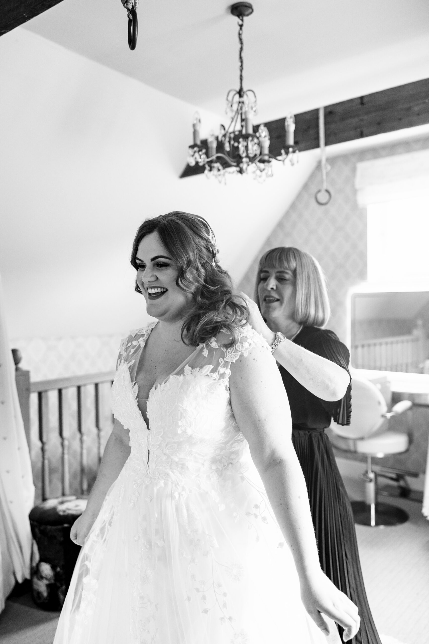 The bride getting in to her wedding dress. She is looking at herself in the mirror and she is very happy. She has a lady helping her do up her back buttons.