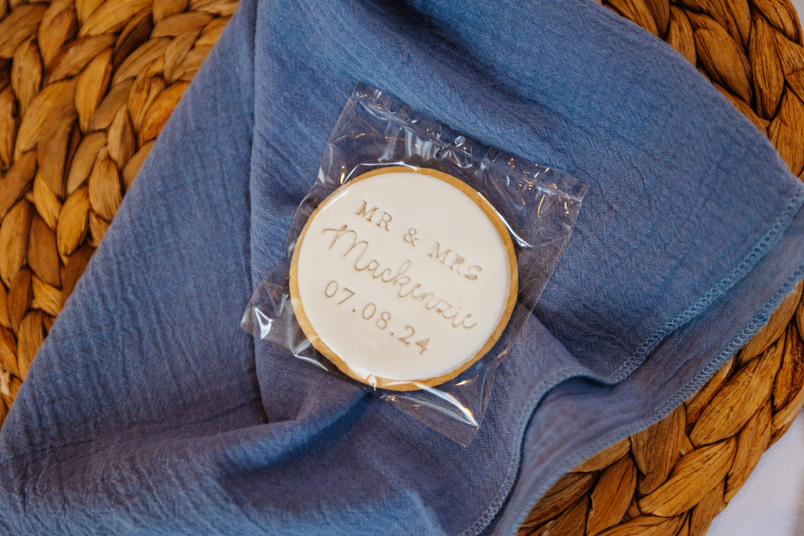 A blue napkin and a biscuit in cellophane on top of it. The biscuit has white icing on top and it has Mrs & Mrs Mackenzie embossed in to it as well as 07.08.24 (the date of their wedding).