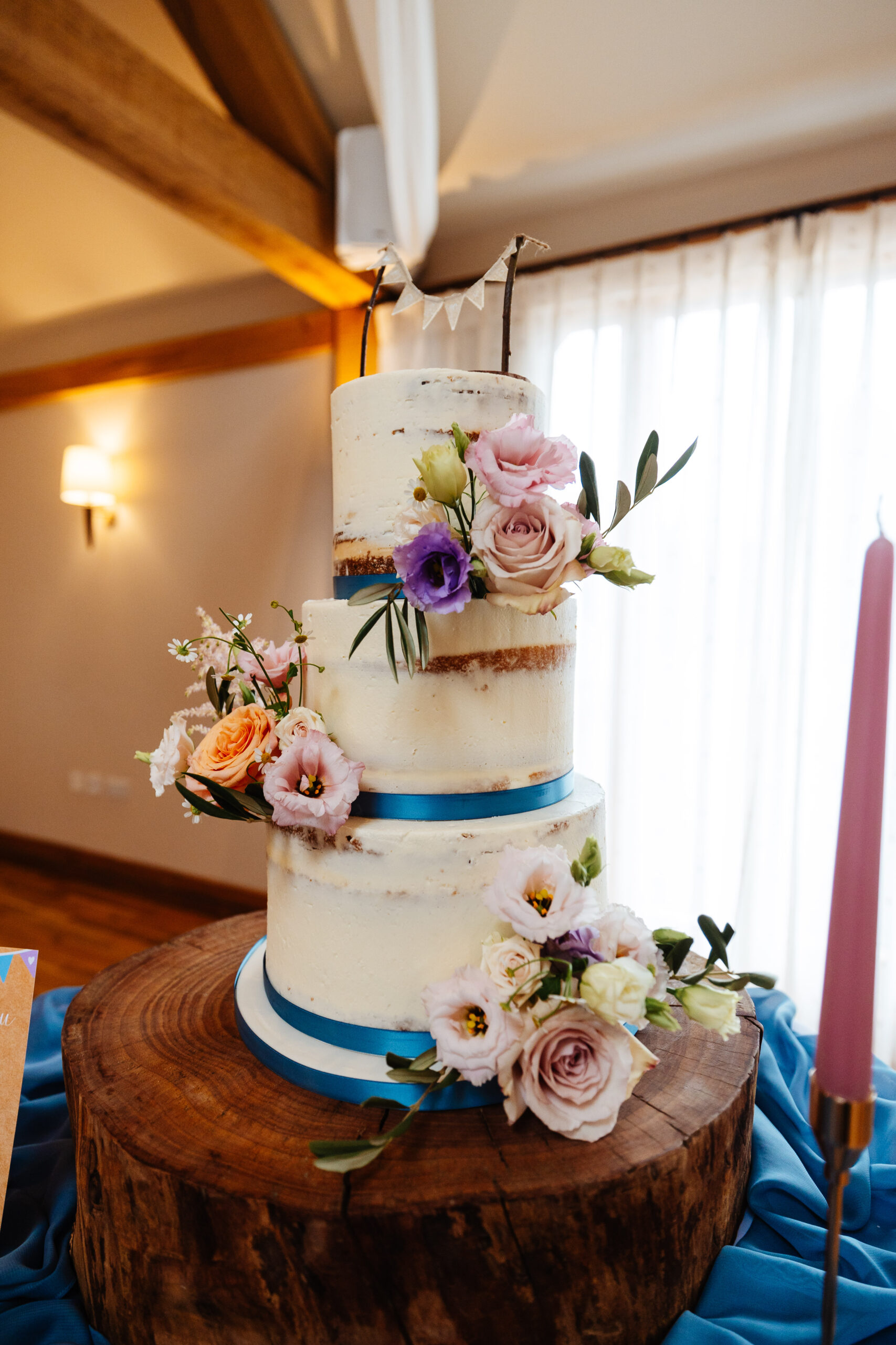 A three tiered cake with blue ribbon tied around the bottom of each layer. It has naked white icing on it and it is decorated with flowers scattered on it.