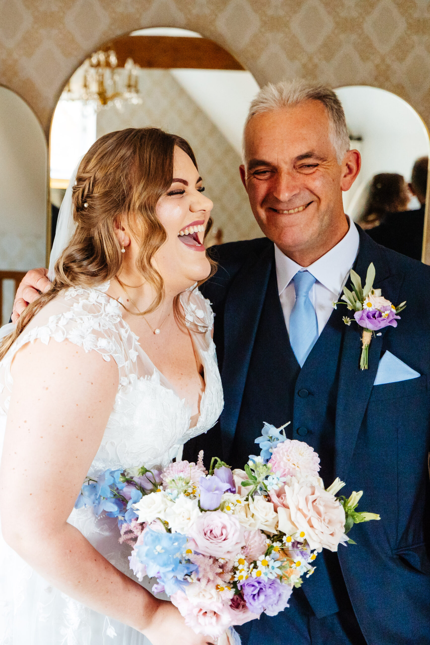 A bride and her father. Her father is in a navy suit and has a little button hole of a purple flower and some daisies. The bride has her mouth wide open laughing and her Dad looks very happy.