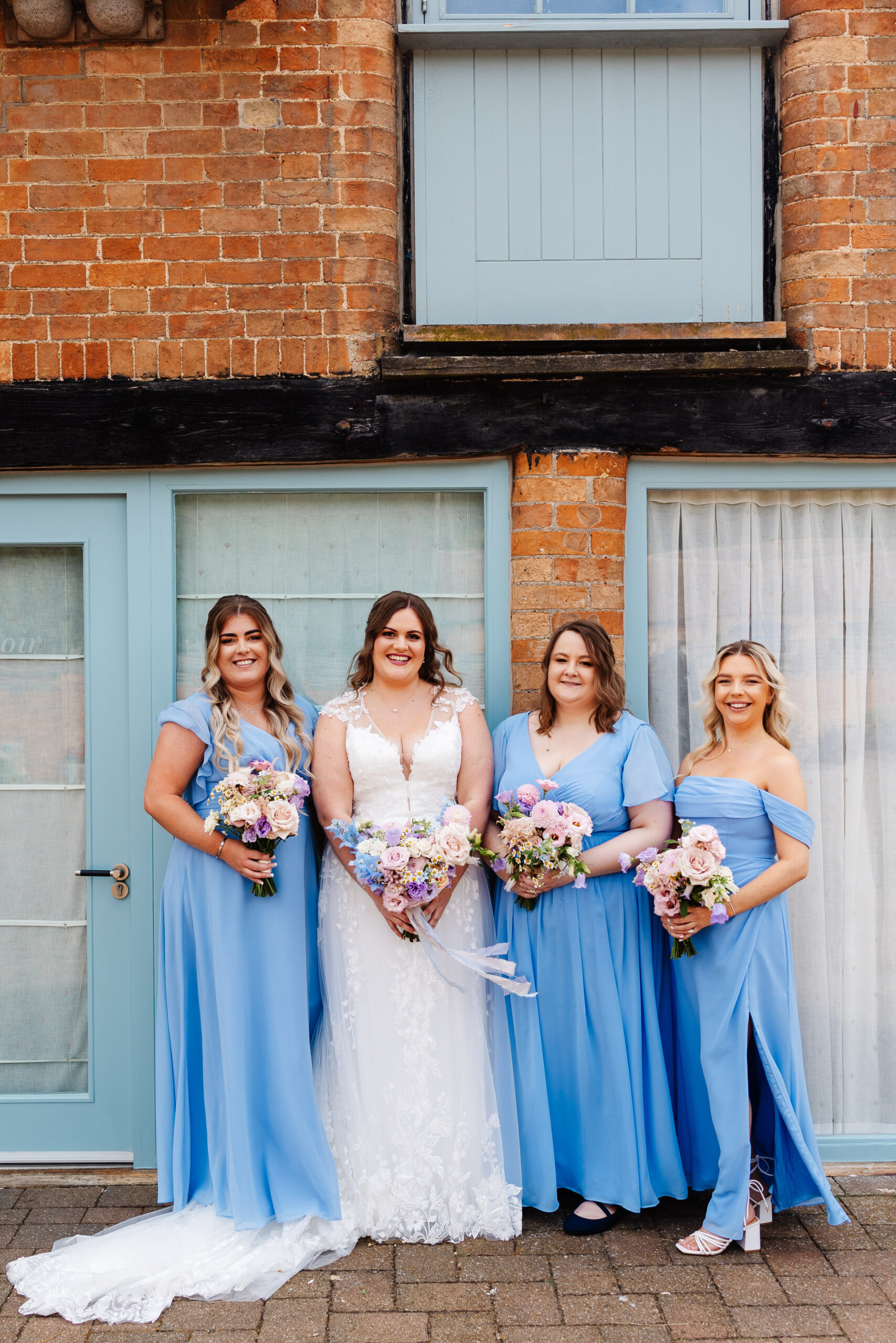 The bride and her bridesmaids outside of Bassmead. The bridesmaids are wearing a variety of style dresses in a pale blue colour. They are all holding bouquets of pale pink roses, dahlias and purple and blue flowers.