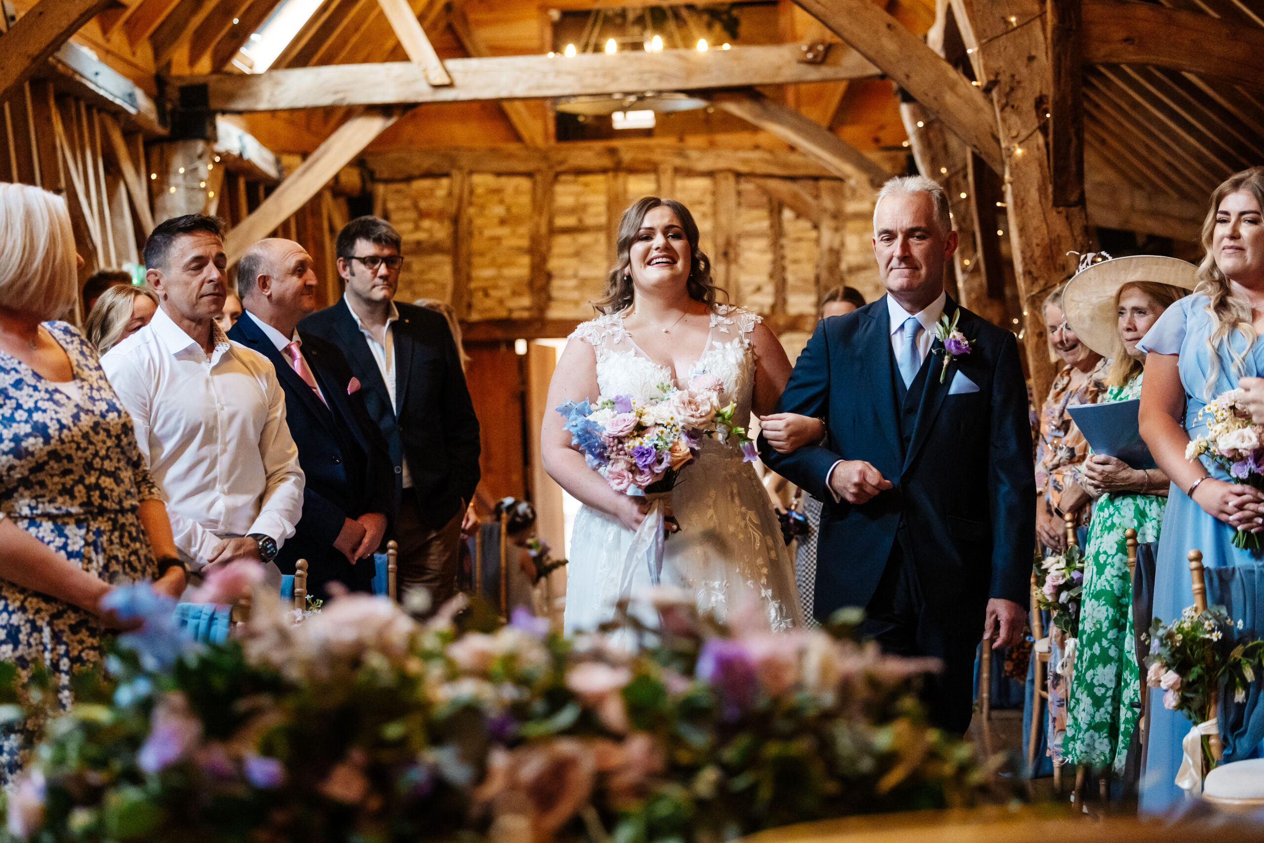 The bride and her father walking down the aisle. She is smiling but looks emotional. The guests around her look emotional too.