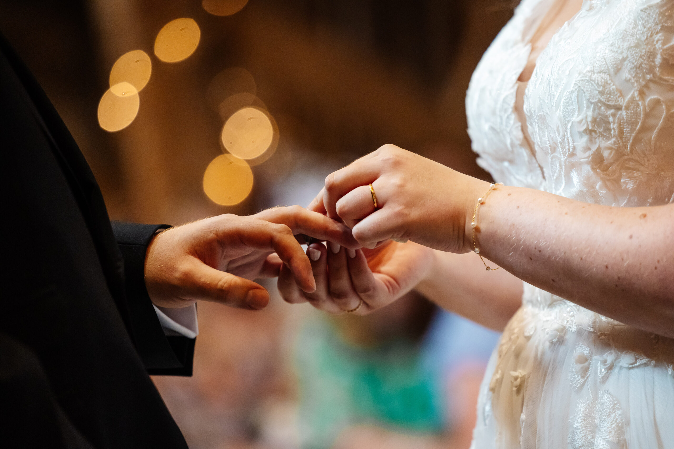 The bride putting a ring on her groom's finger. The light in the background is soft.