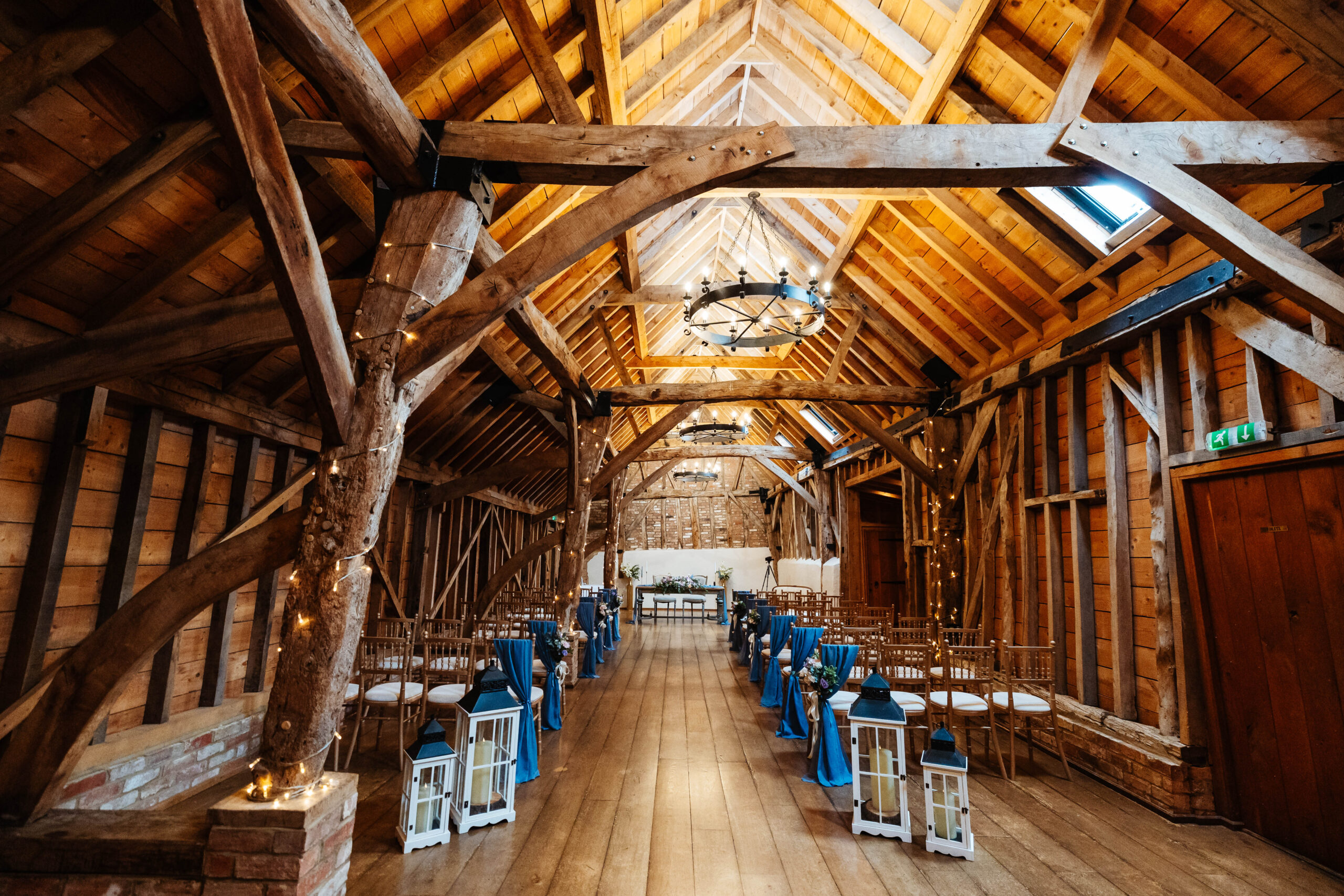 The barn venue ready for the couple to be married in. There are blue drapes on the end of each chair and some fairy lights dotted around the wooden beams.