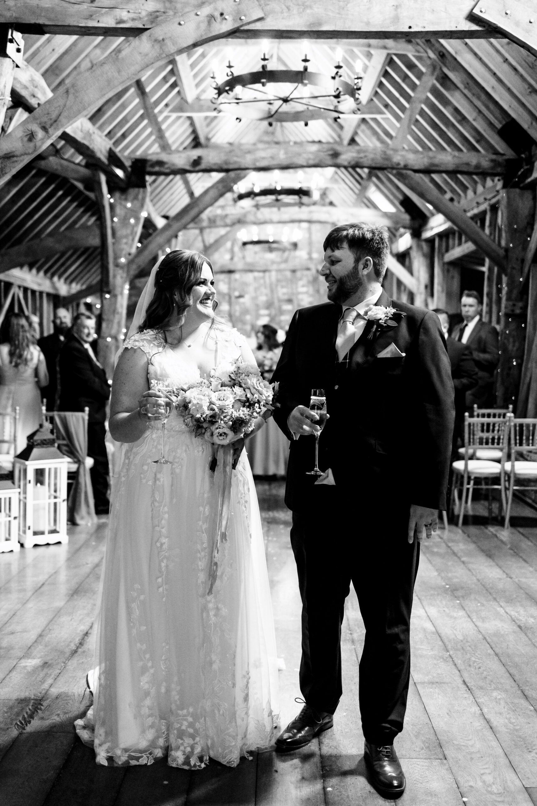 A black and white image of the bride and groom. They are holding a glass of champagne and looking at each other. You can see happy guests in the background.