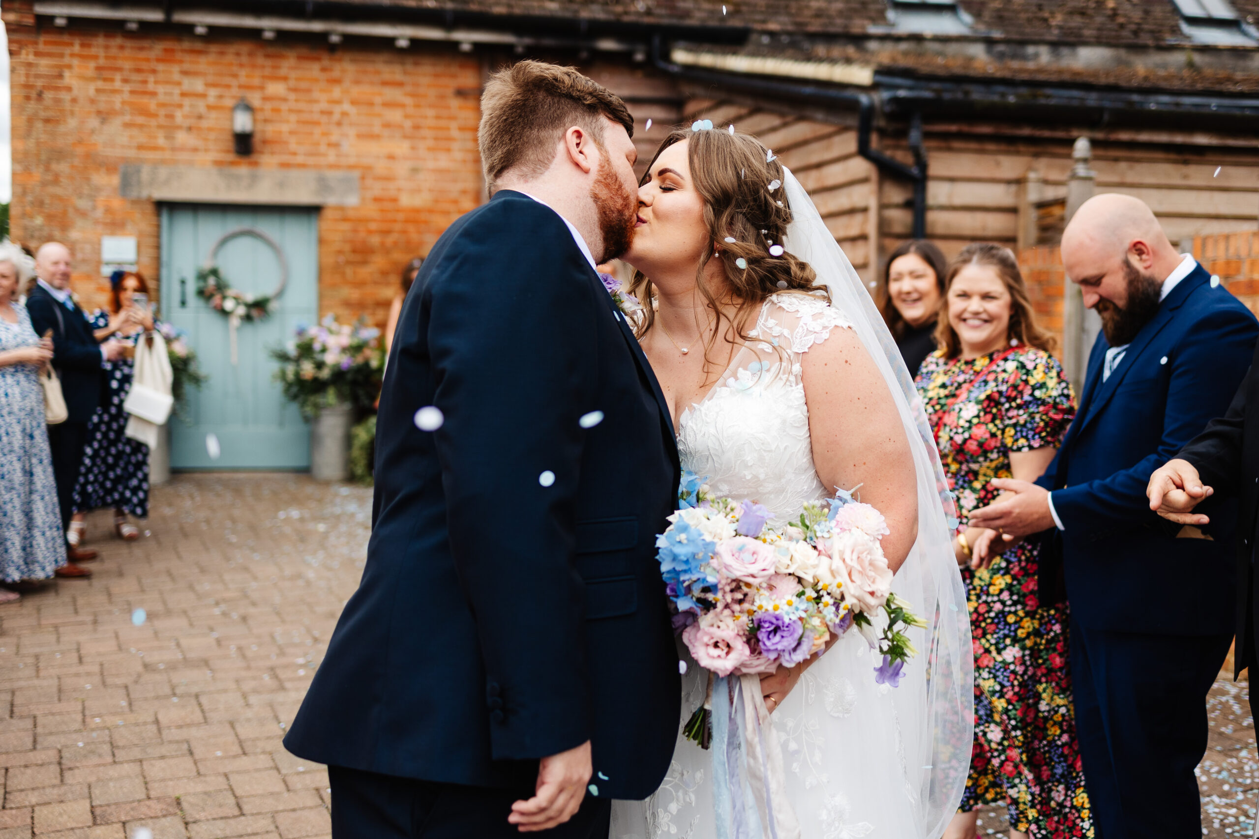 The bride and groom kissing. They have had confetti thrown around them and it is in the air and in their hair. The guests in the background are smiling.