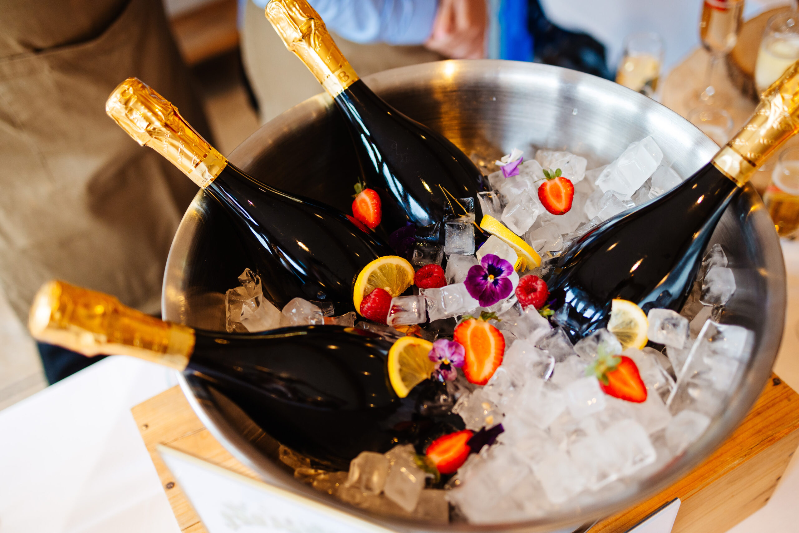 Four bottles of champagne on ice. There are strawberries, lemon slices, raspberries and edible flowers on the ice too.