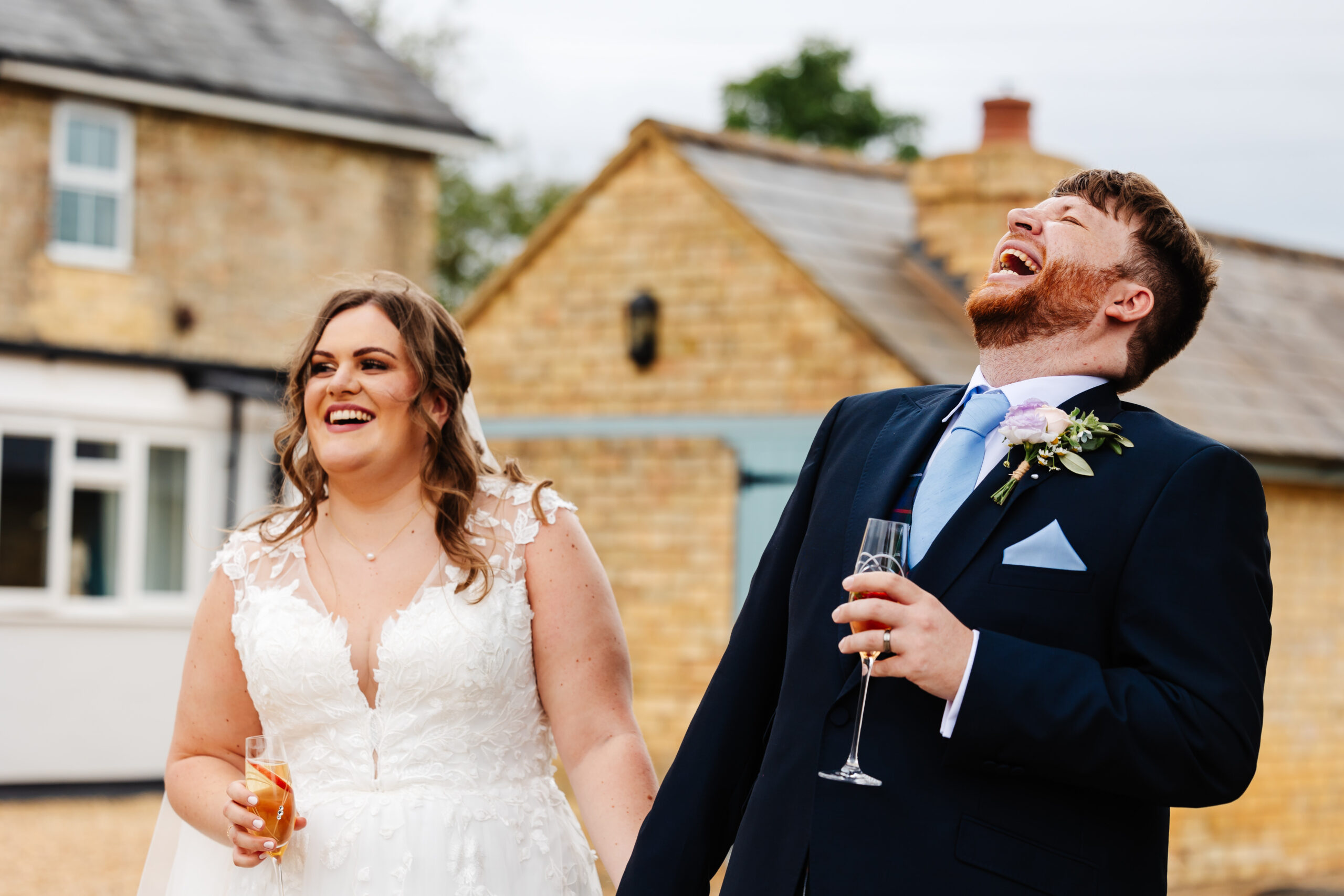 A candid photo of the bride and groom outside smiling. The groom is throwing his head back in laughter.