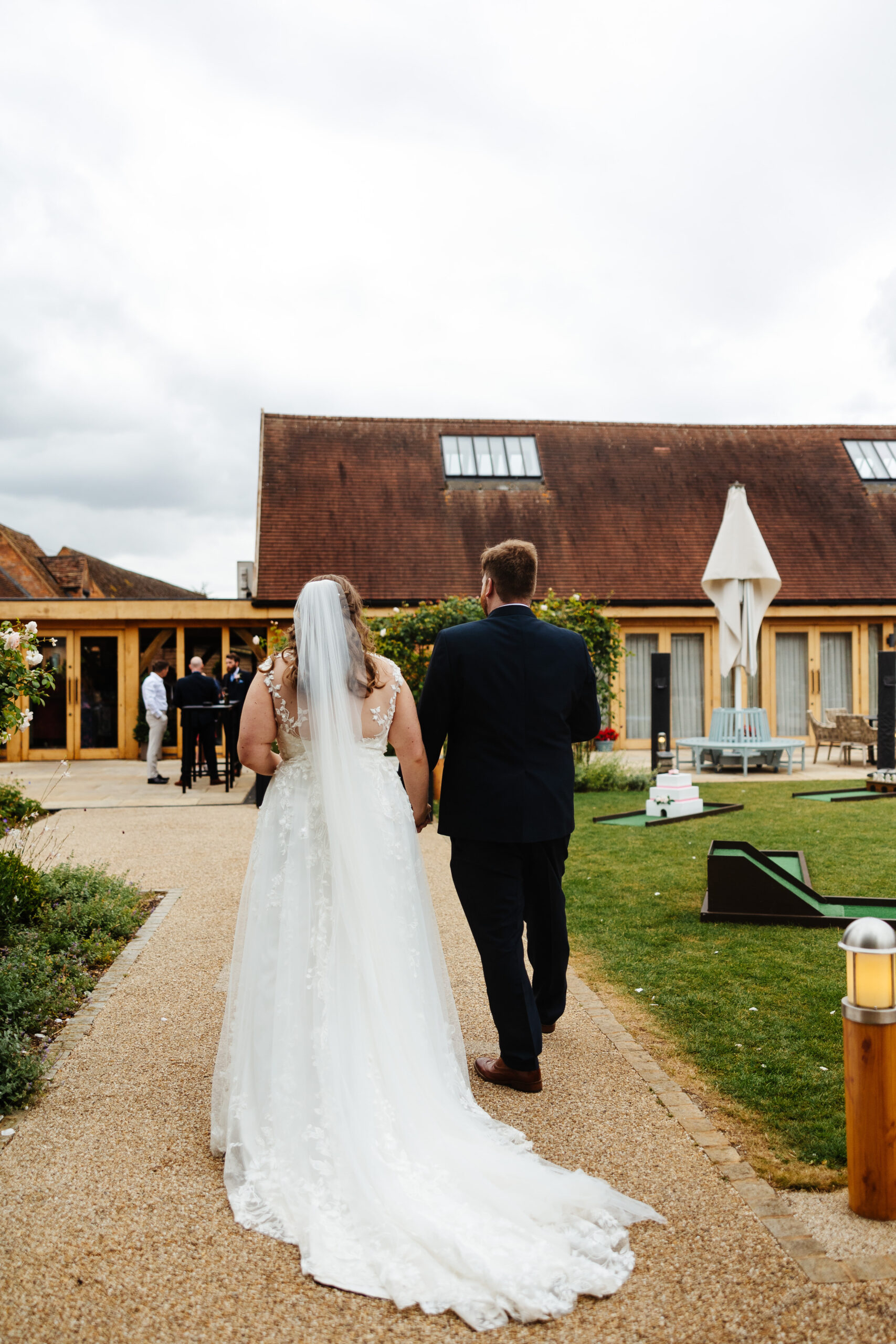 The back of the bride and groom as they are walking towards their guests. The bride's dress has a long train and a long veil. You can see guests in the background and also mini golf on the grass.