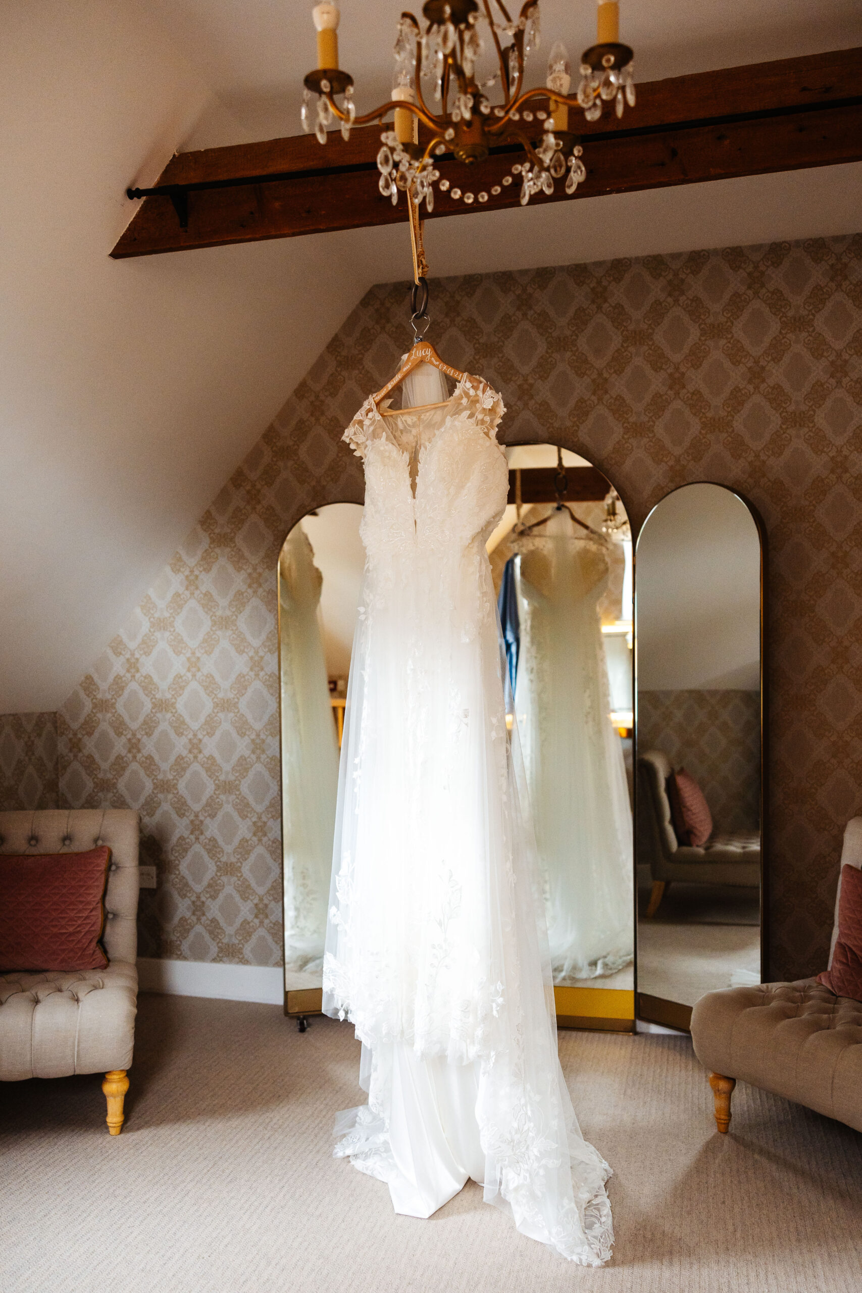 The bride's dress hanging up on a coat hanger from the wooden beam. It is sleeveless with lace and sheer material on the shoulders. It has a long train which has lace detail.