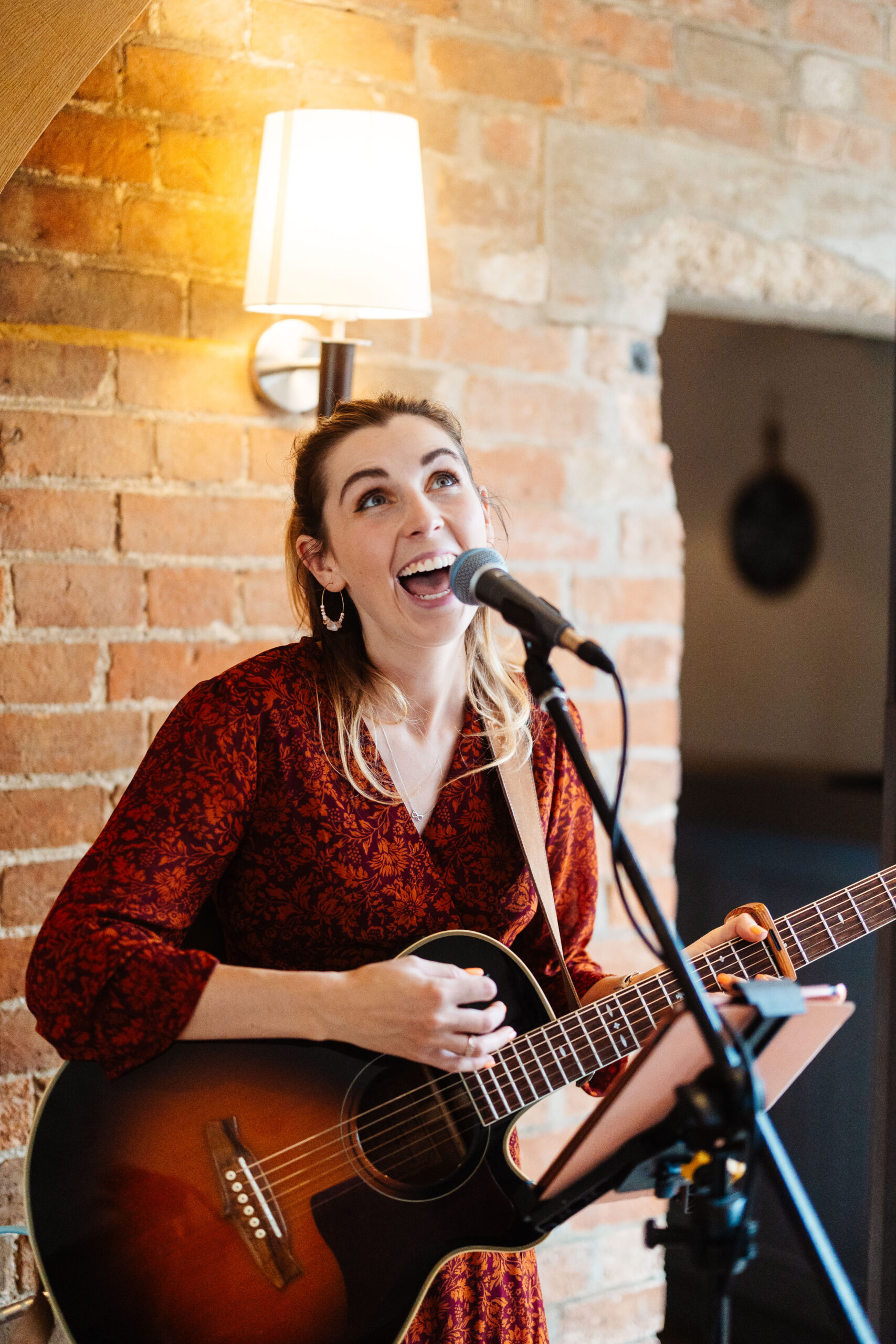 A photo of a musician. She is wearing a red dress and holding a guitar and is singing in to a microphone.