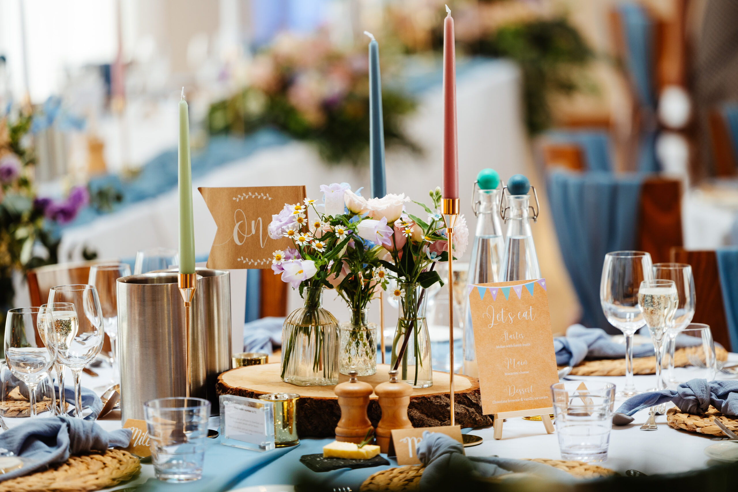 The table centres for the wedding breakfast. They have pastel coloured candles in tall, Gold holders and there is a log slice on the table which has small jars of flowers that match the bride's bouquet. 