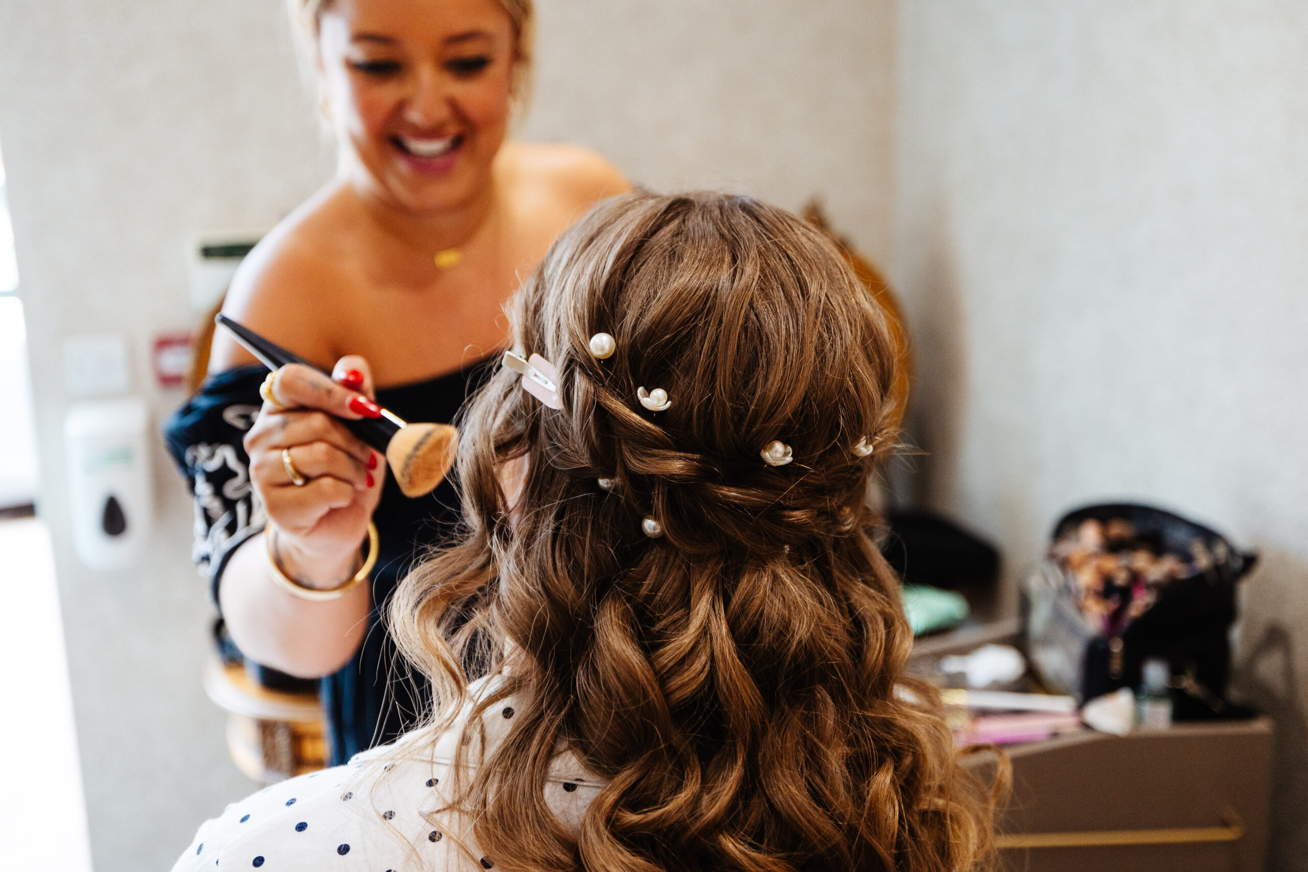 You can see the back of the bride's hair. She has curly hair with a plait across the top half of her head and it has little pearls placed in nicely throughout. You can see the make up artist and she has a blusher brush held closely to Lucy's face. The make up artist is smiling.