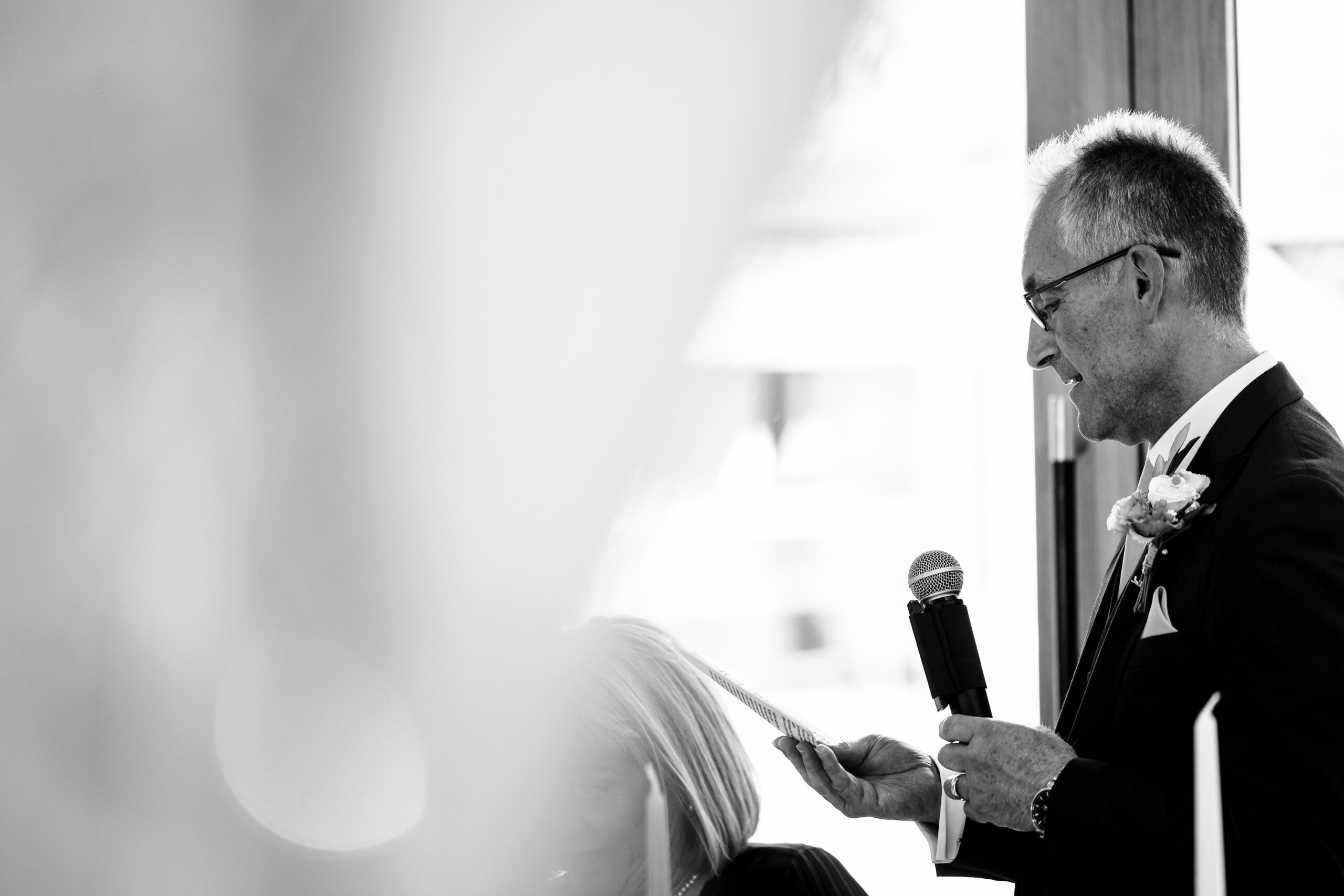 A black and white image of the bride's father reading a speech. He is reading form a piece of paper and holding a microphone.