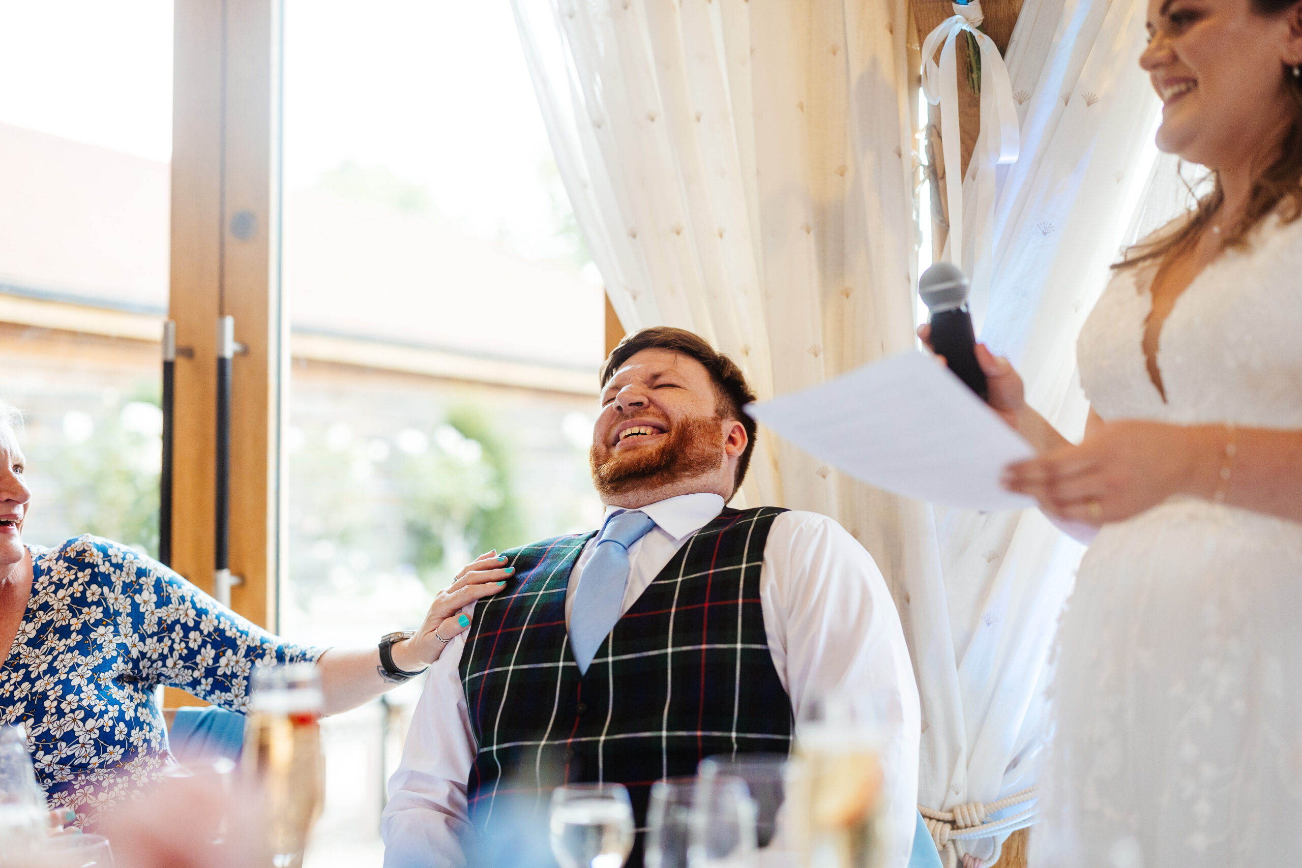 The bride holding a microphone as she reads her speech from a piece of paper. The groom is laughing with his eyes shut and his mum has her hand on his shoulder and is also laughing.