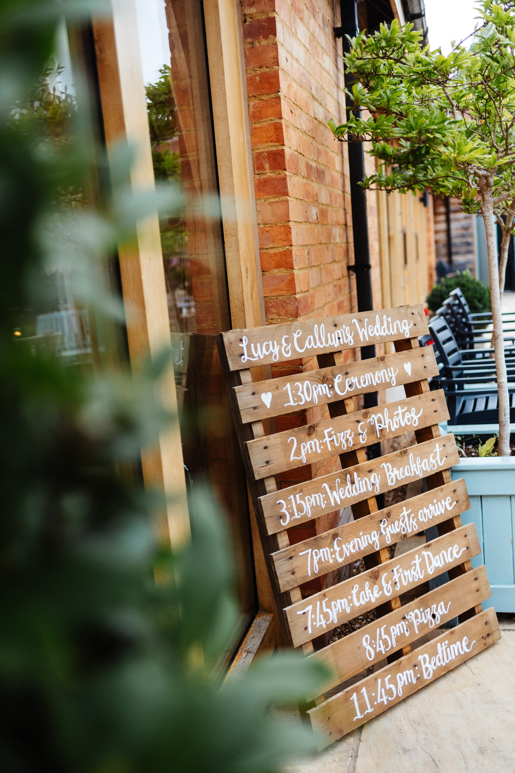 A pallet with the couple's itinerary of the day written on it in white, calligraphy font. It states the times from the ceremony, to fizz and photos, to the wedding breakfast, evening guests arriving, cake and the first dance, pizza and then finally, bedtime.