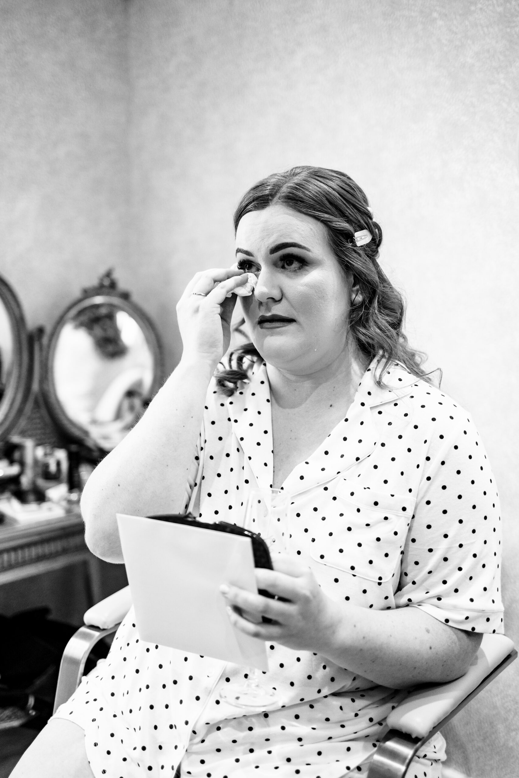 A black and white image of the bride. She is in black and white spotted pyjamas and she is reading a letter from a loved one. She is crying and holding a tissue to her eye.