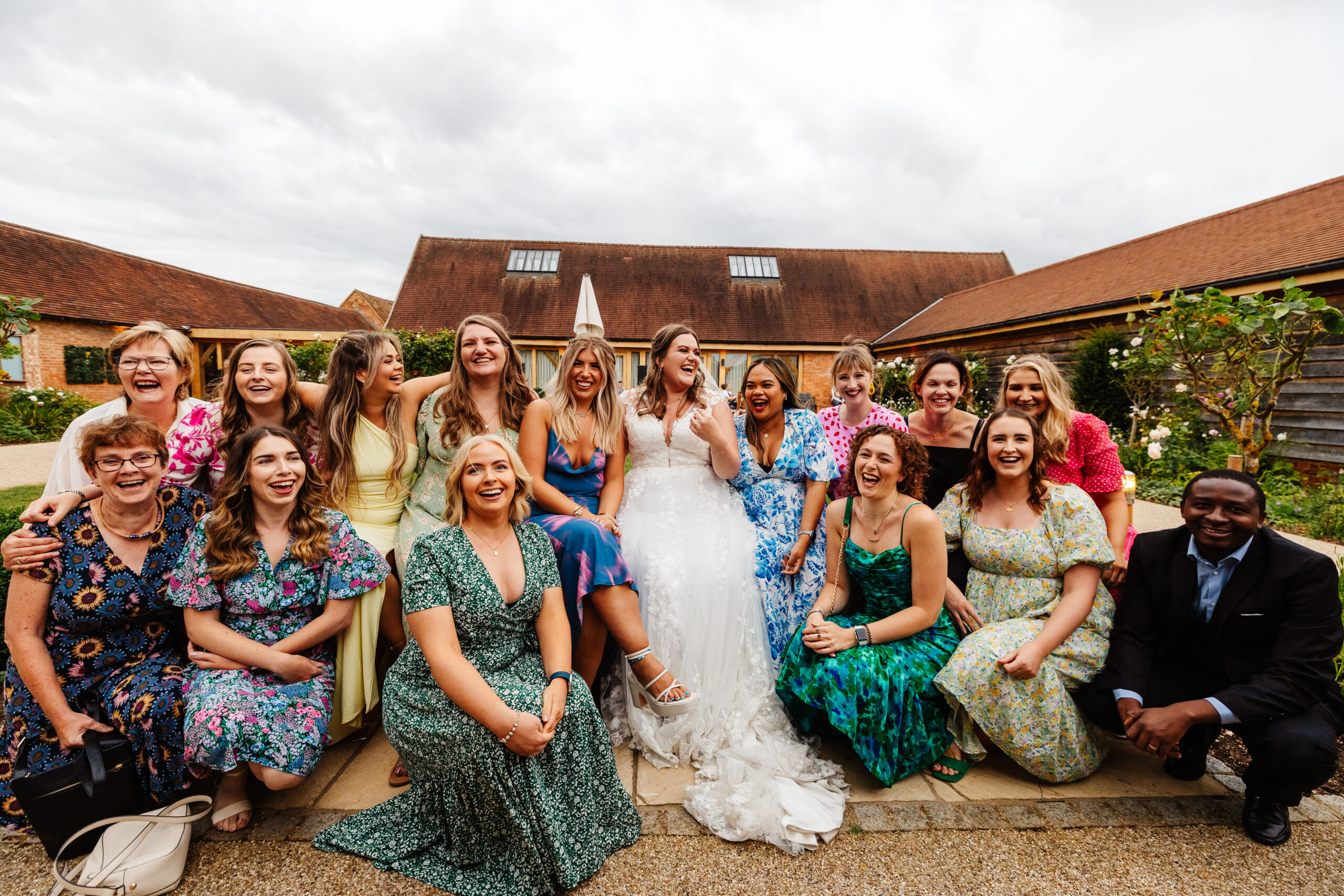 The bride and a group of her friends. They are outside sat down in a clump and on the floor and are all smiling and laughing.