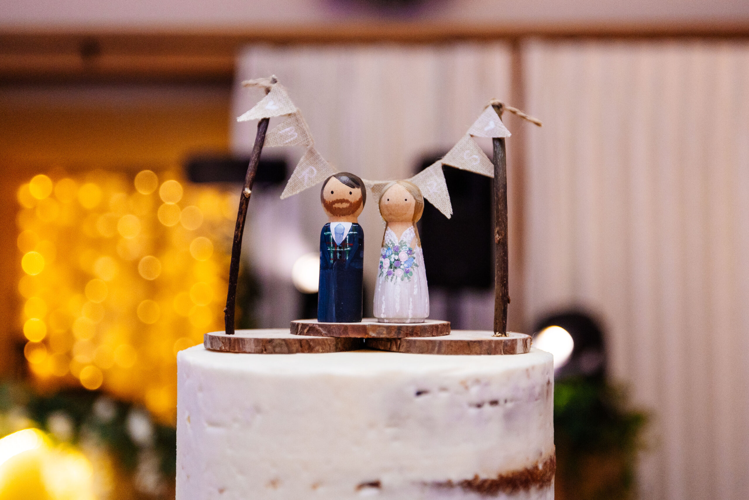 A photo of the top of the cake. It has a two wooden figures of the bride and groom with two sticks and bunting over the top that says Mr & Mrs.