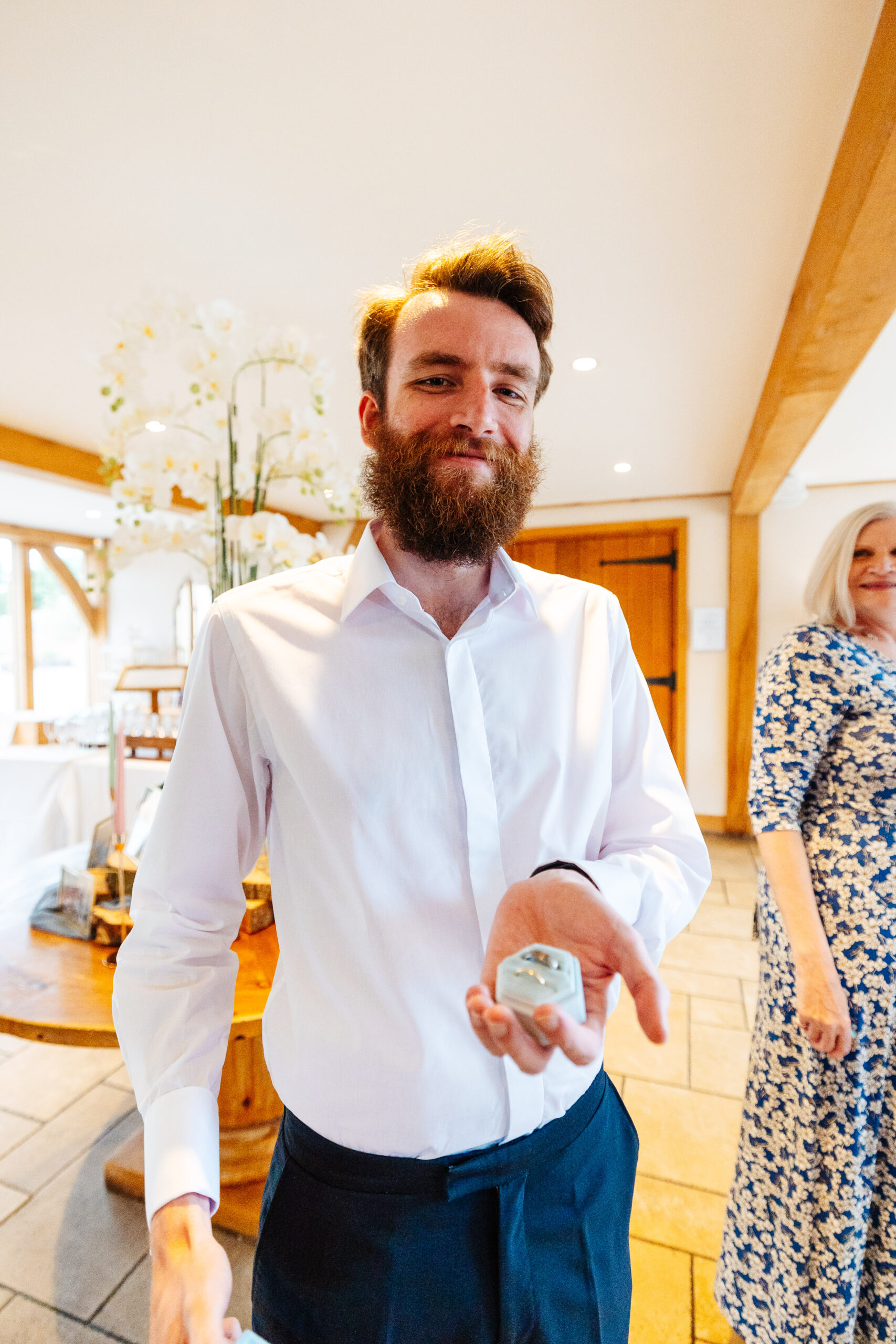 One of the groom's party. He is wearing a white shirt with the top button undone and he is holding the ring box which is a pale blue, hexagonal shape box. It has two rings inside it. He is smiling.