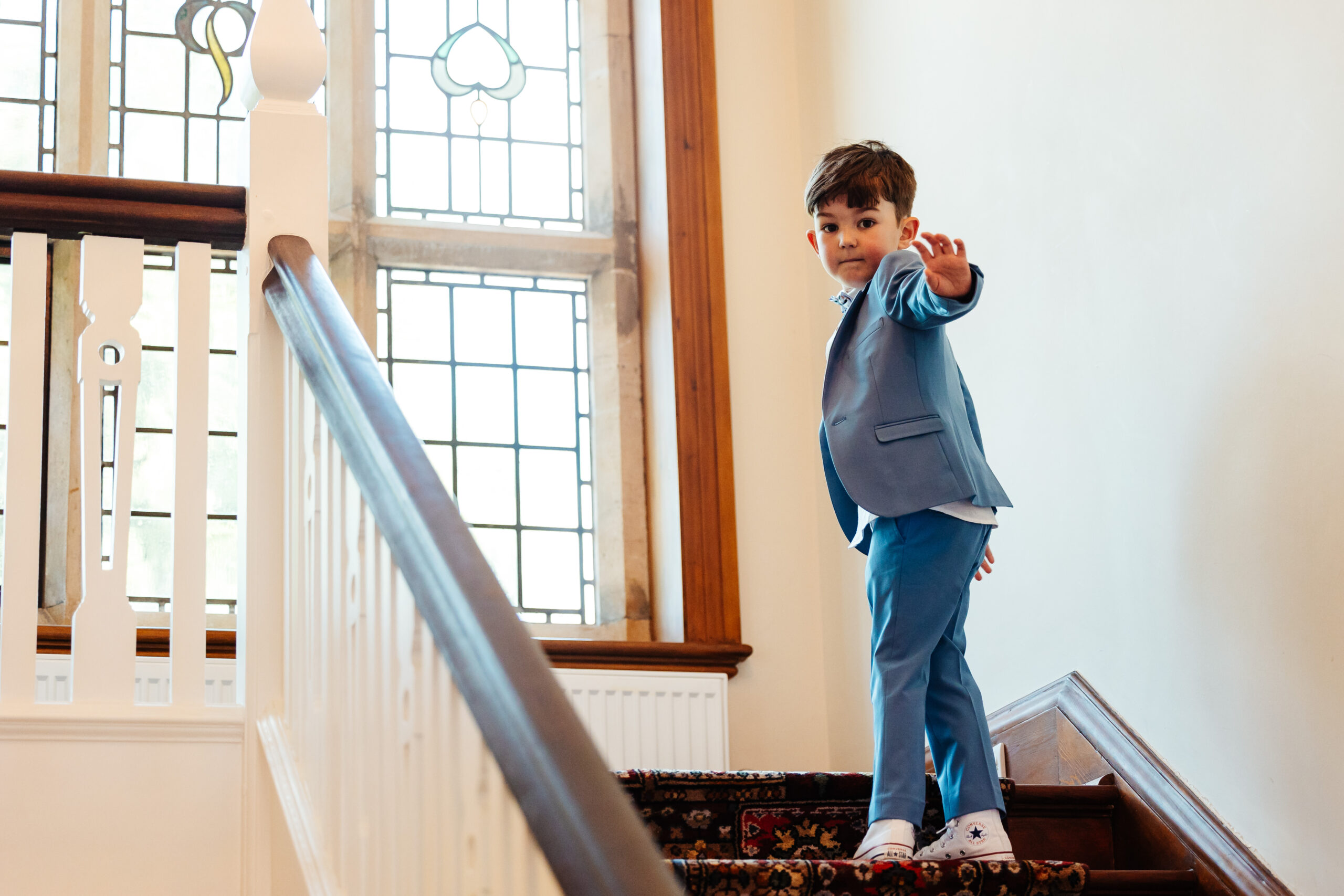 A little boy in a pale navy suit and white converse trainers. He is standing at the top of the stairs and looking back at the camera waving.