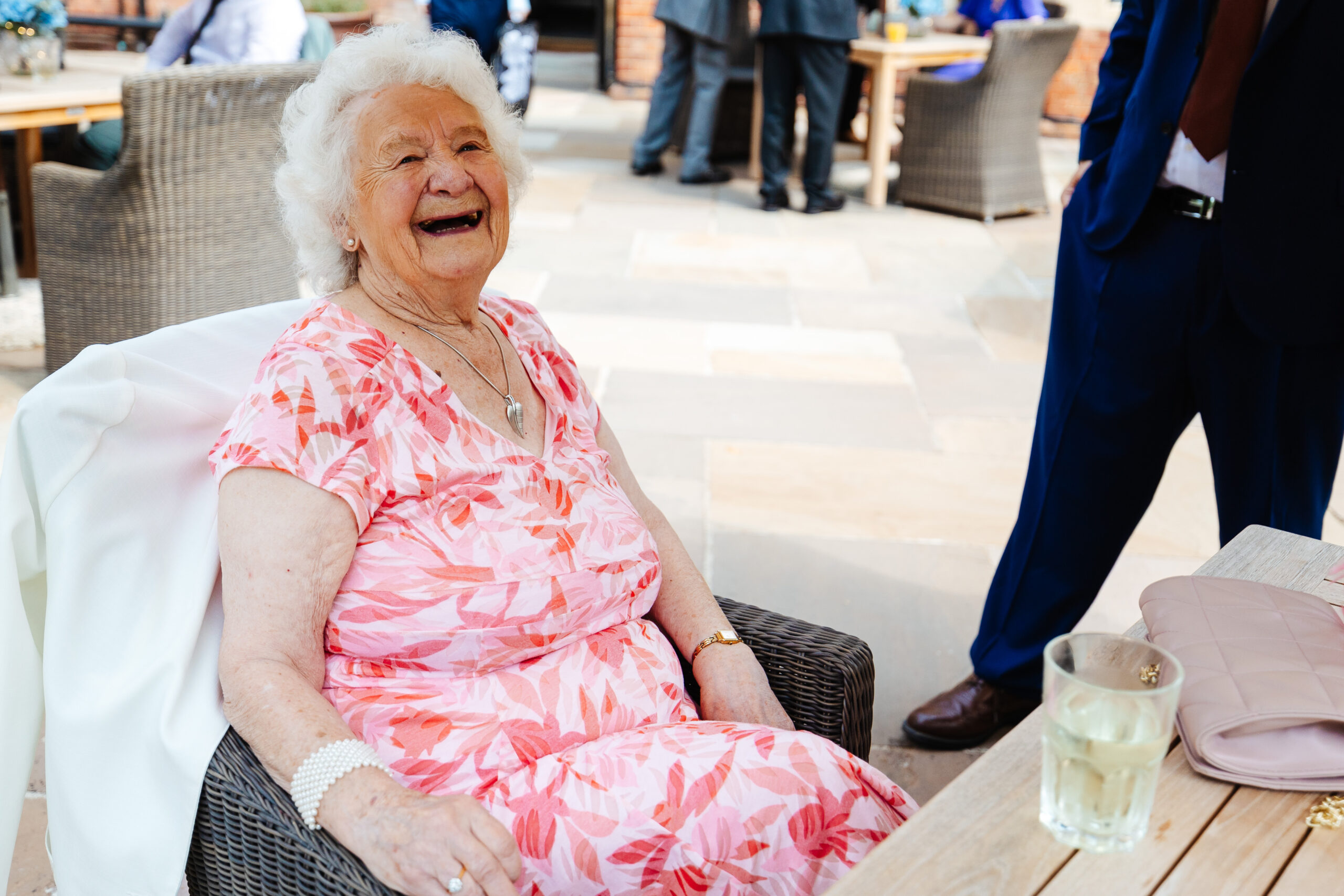 Nan sitting down having a drink and smiling. She is in a pale pink dress with floral designs on.