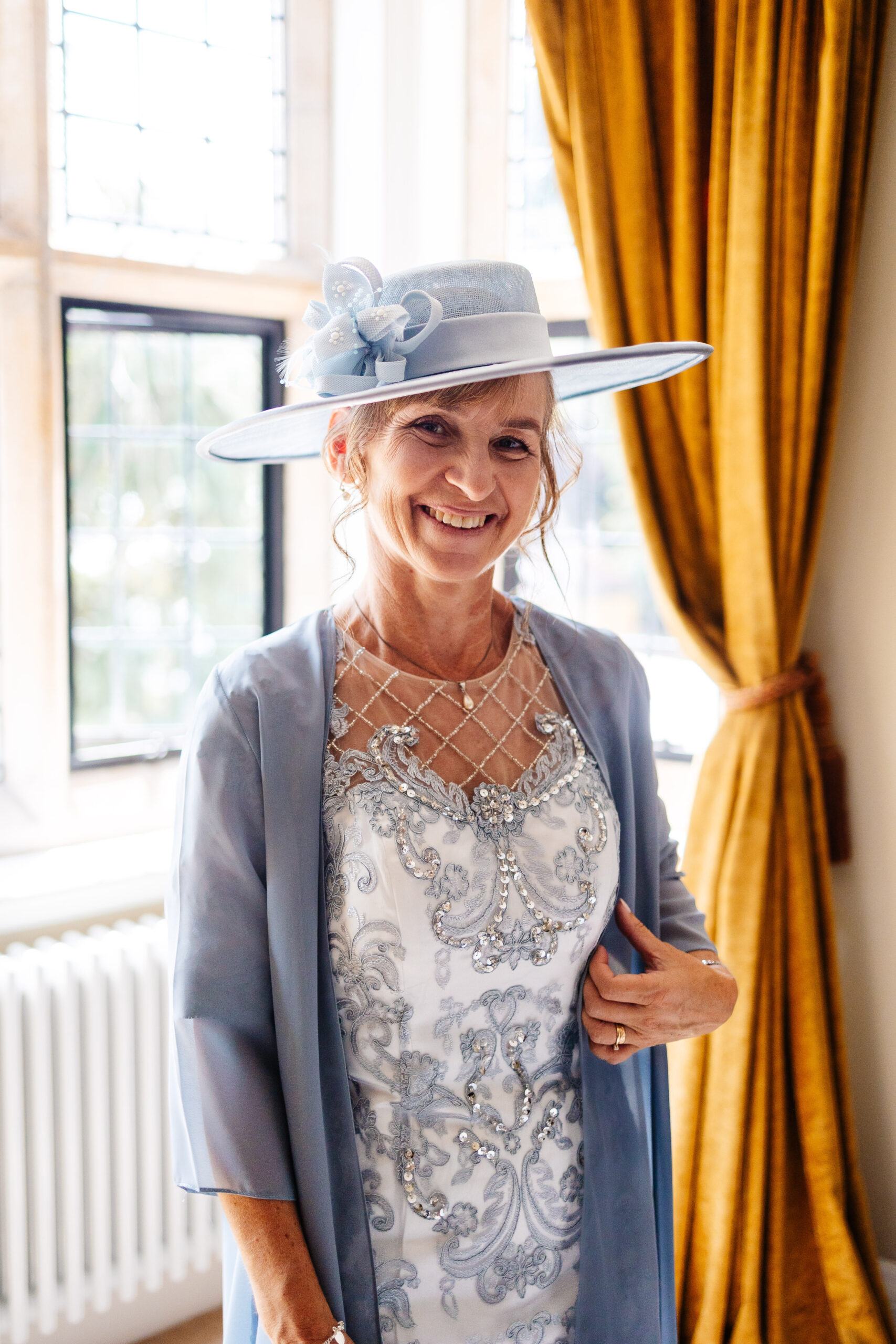 The mother of the bride. She is wearing a pale blue/grey dress with lots of lace detailing and beaded on the front and a grey blazer with a large grey hat. She is smiling at the camera.