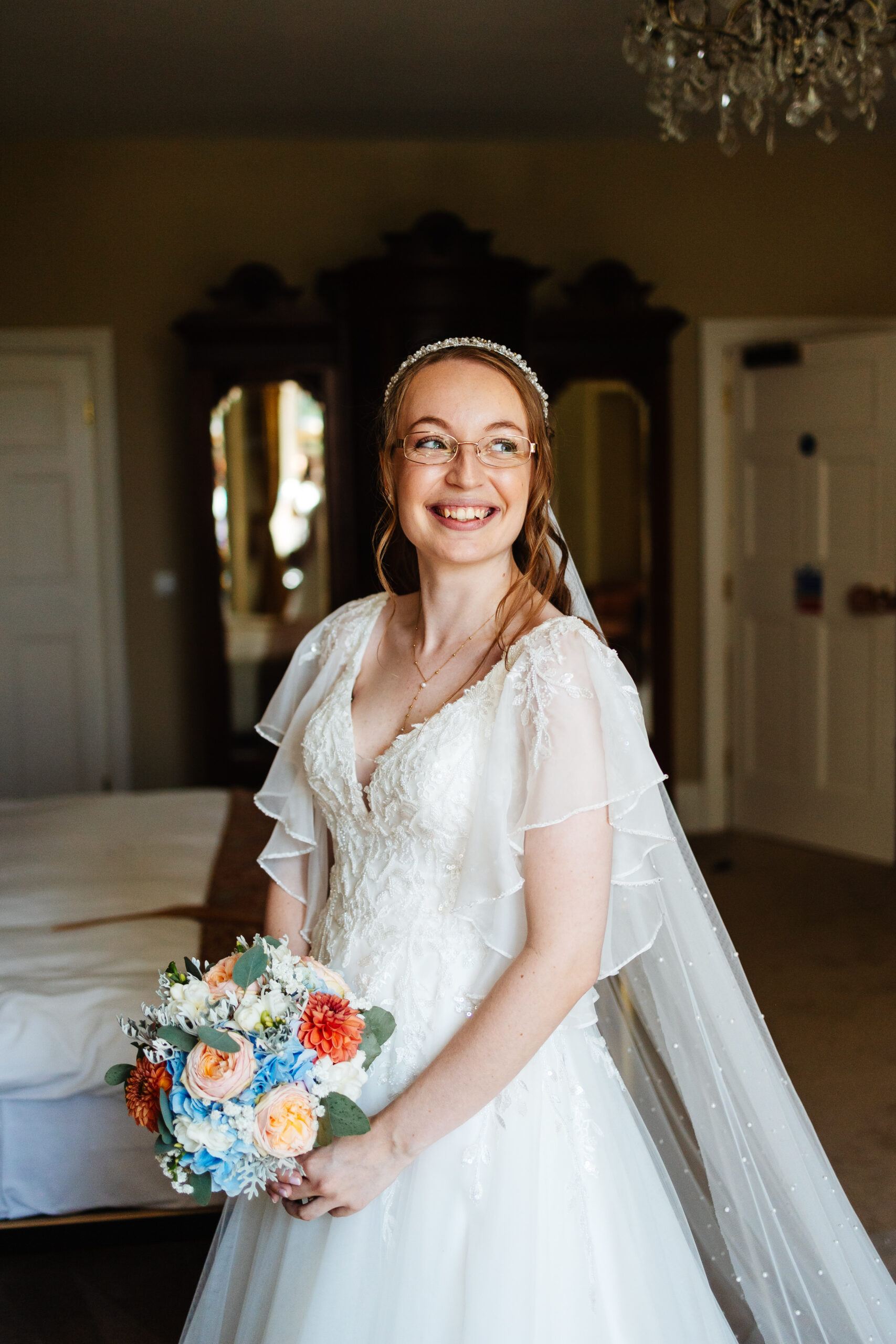 The bride in all her glory. She is in her dress and veil and is holding her bouquet. She is looking away from the camera and smiling.