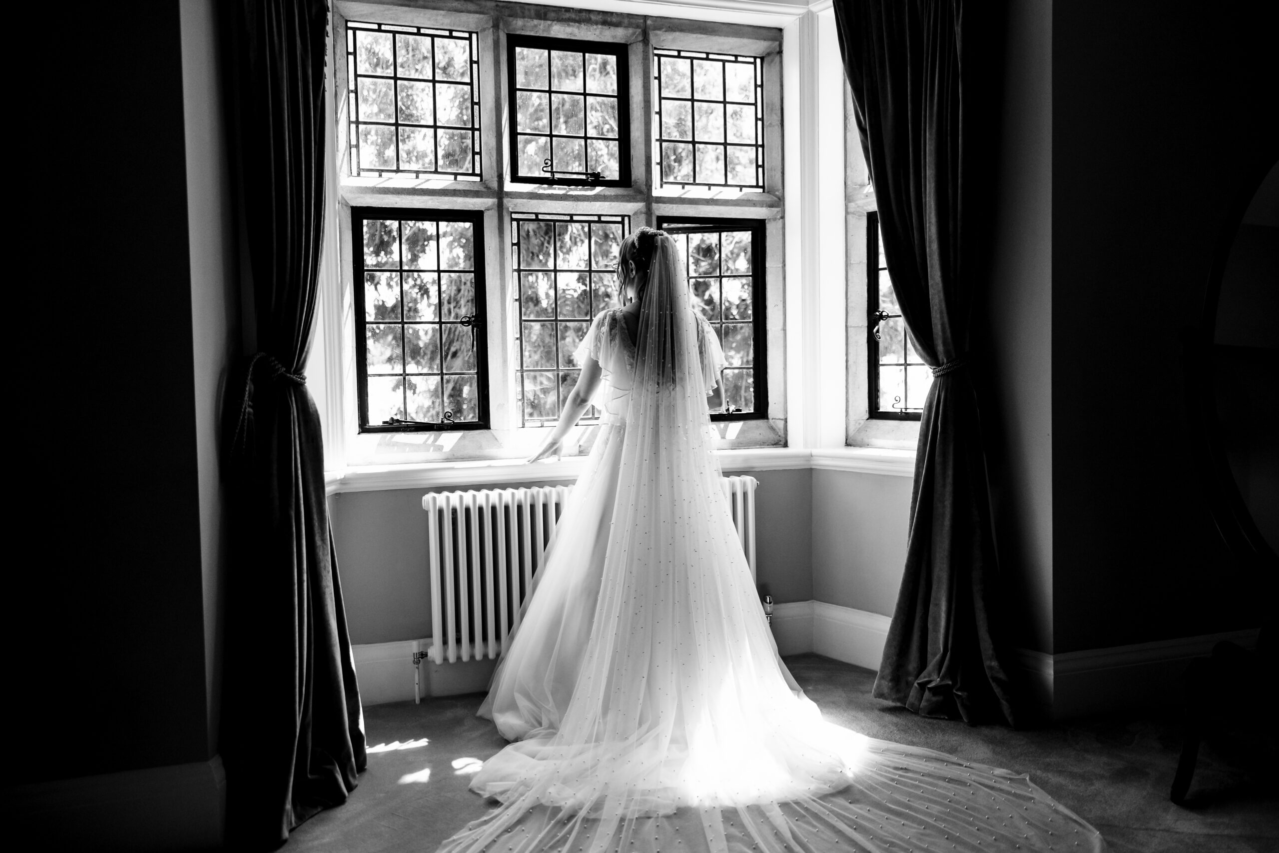 A black and white image of the bride. She is looking out of a window and you can see the flow of her veil and train of her dress spread out across the floor.