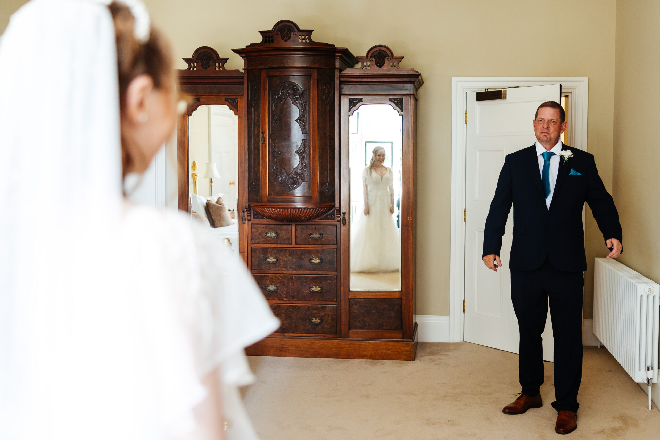 The father of the bride walking in to see his daughter in her wedding dress for the first time. You can see the bride in the mirror and she is smiling and the father looks emotional.