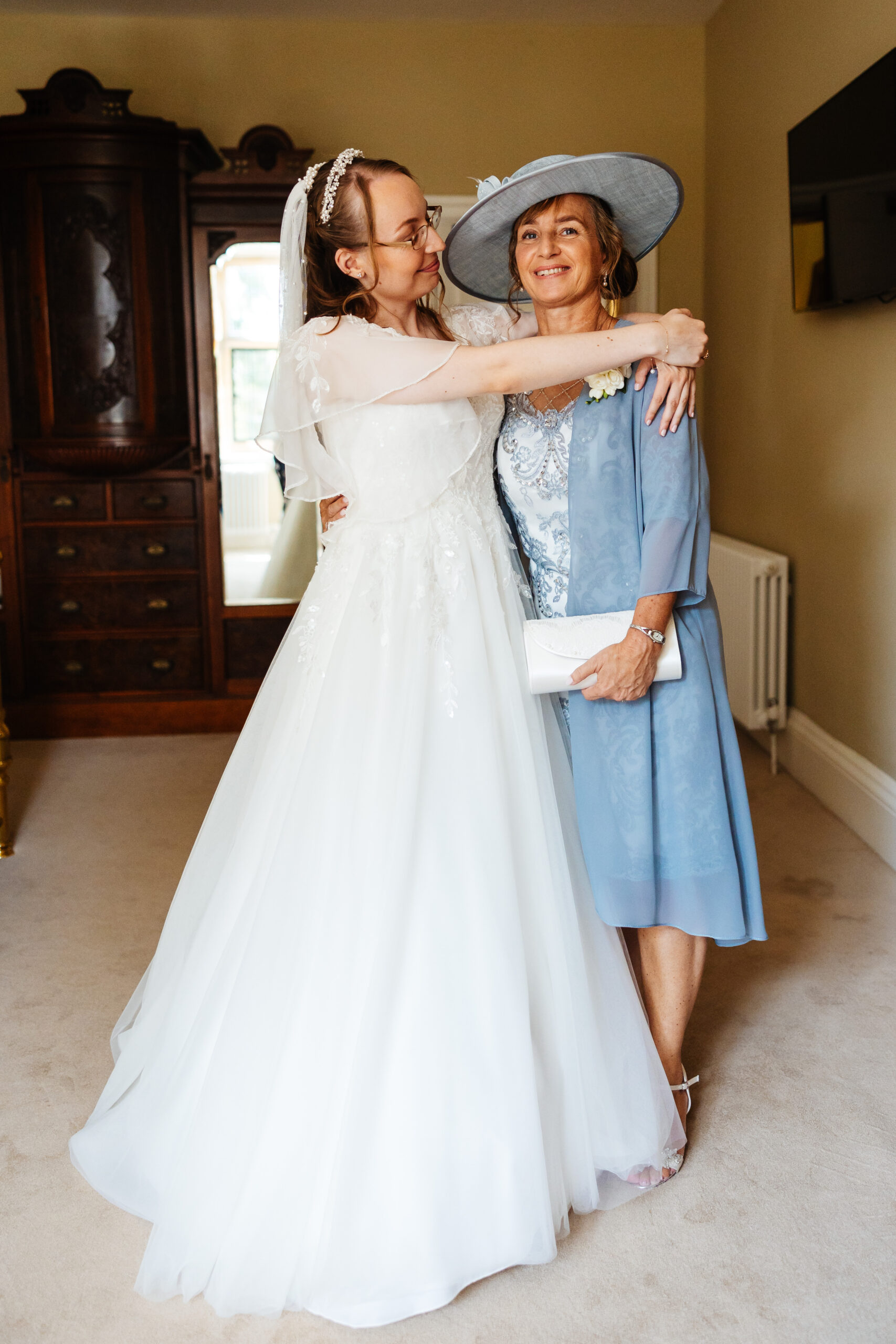 The bride and her mother. The bride is looking at her mum and smiling and the mother is looking at the camera and smiling.