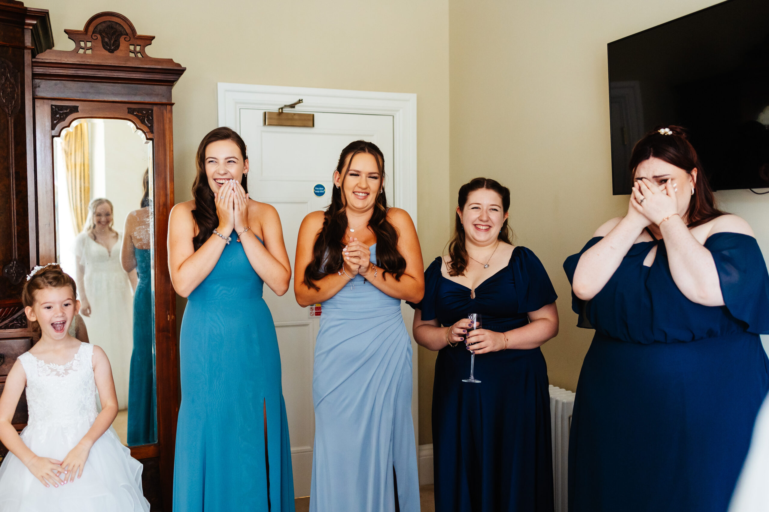 The bridesmaids first look of the bride in her dress. There are 4 bridesmaids and one flower girl. The girls all have happy smiling faces and one has her hands over her mouth and is crying. The flower girl has her mouth open and looks really proud.