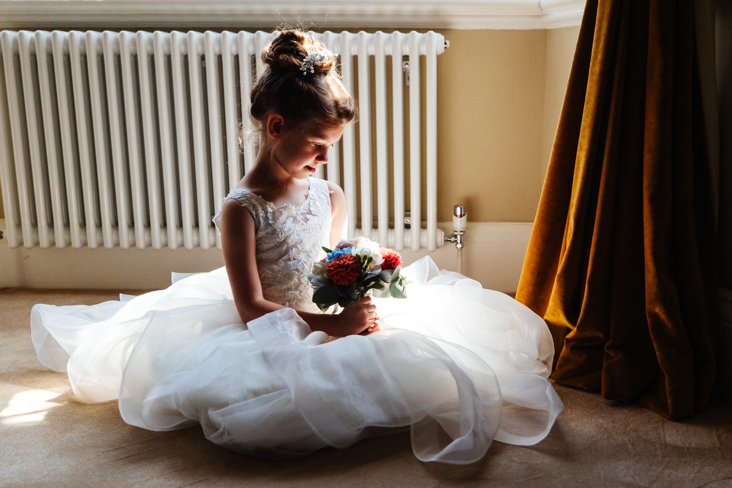 The flower girl sat down looking at her bouquet of flowers. She has her hair tied up with some little diamonds in it and her dress is flowing and white with a laced top.