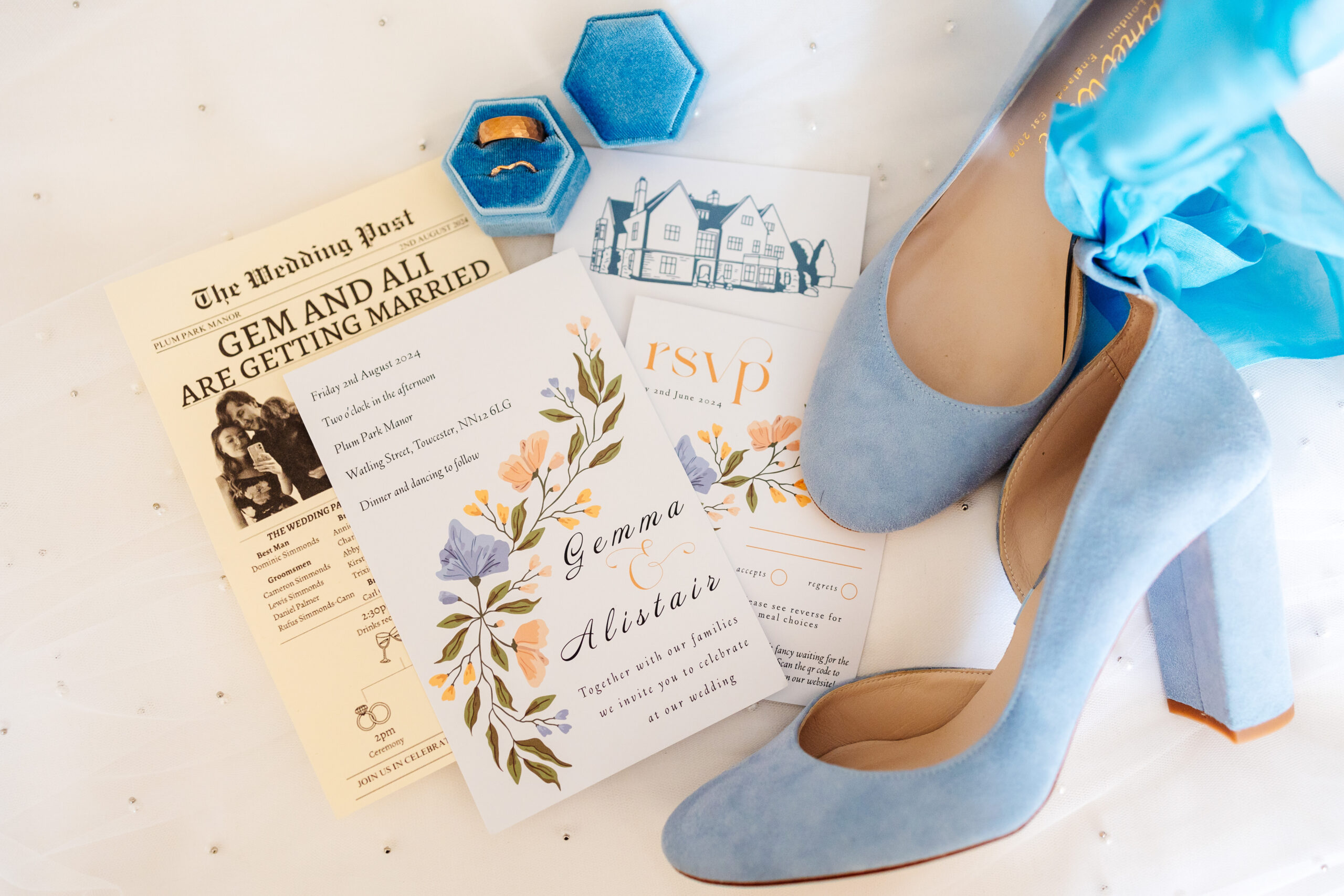 A flat lay image containing lots of details of the wedding including the bride's pale blue, suede shoes, an invite and the ring box containing the wedding rings.