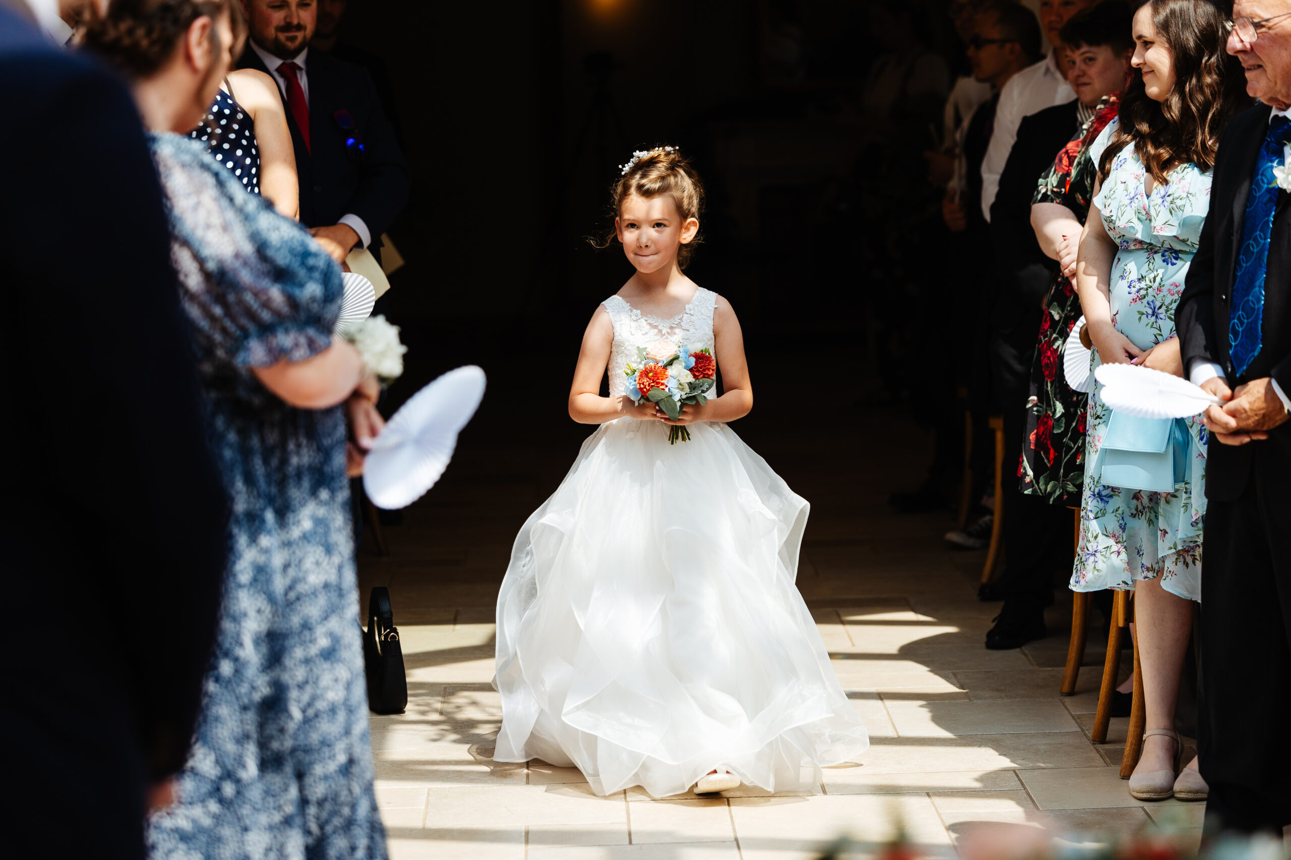 The flower girl walking down the ailse. She is holding a bouquet of bright flowers and looks happy. All guests are looking at her and they are smiling.