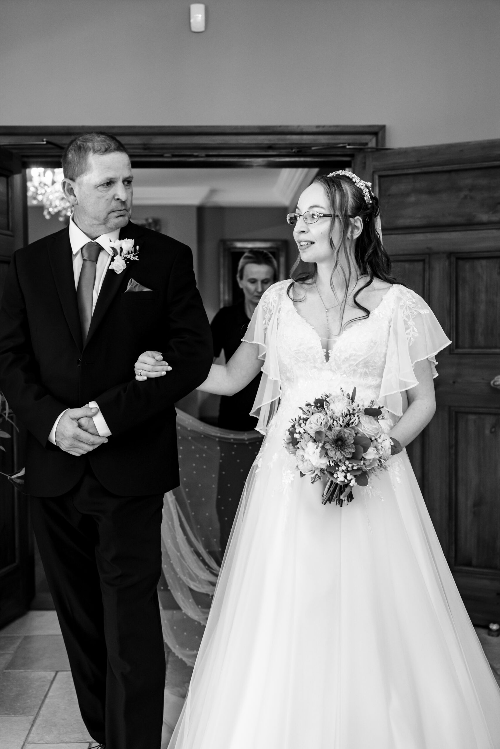 A black and white image of the bride walking down the aisle. She is holding her dad's arm and is looking a little nervous.