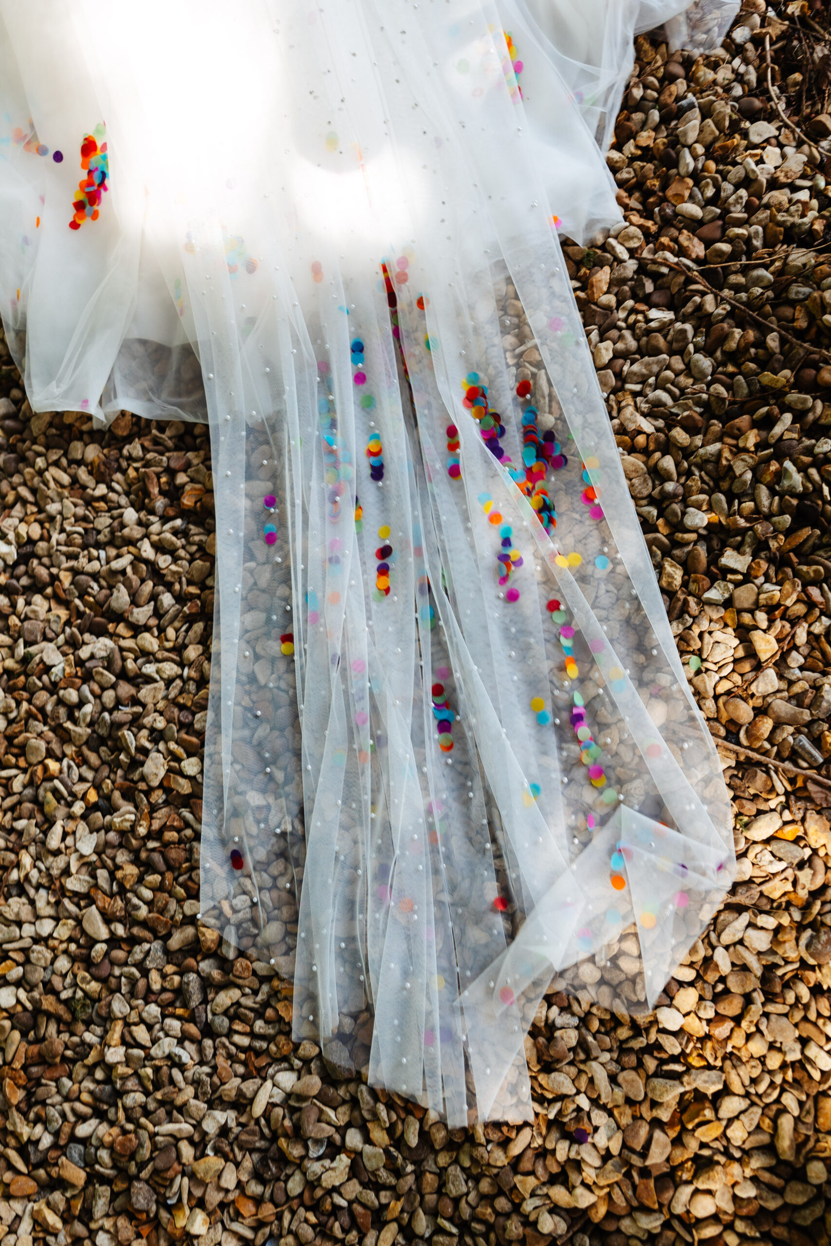 The bride's veil on the floor on gravel. There is bright confetti on her veil. 