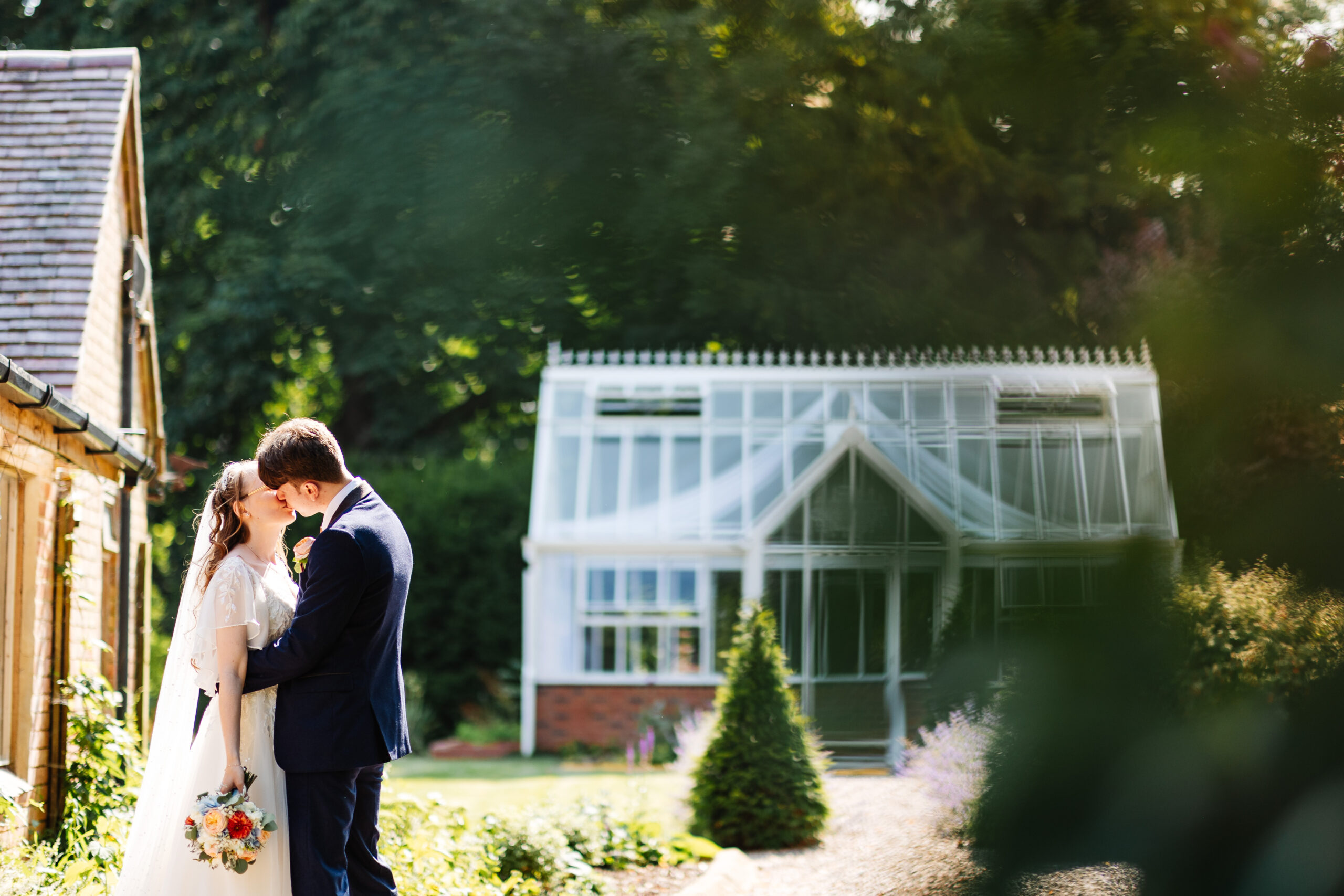 The bride and groom outside kissing. You can see the beautiful trees in the background and the sun is shining.