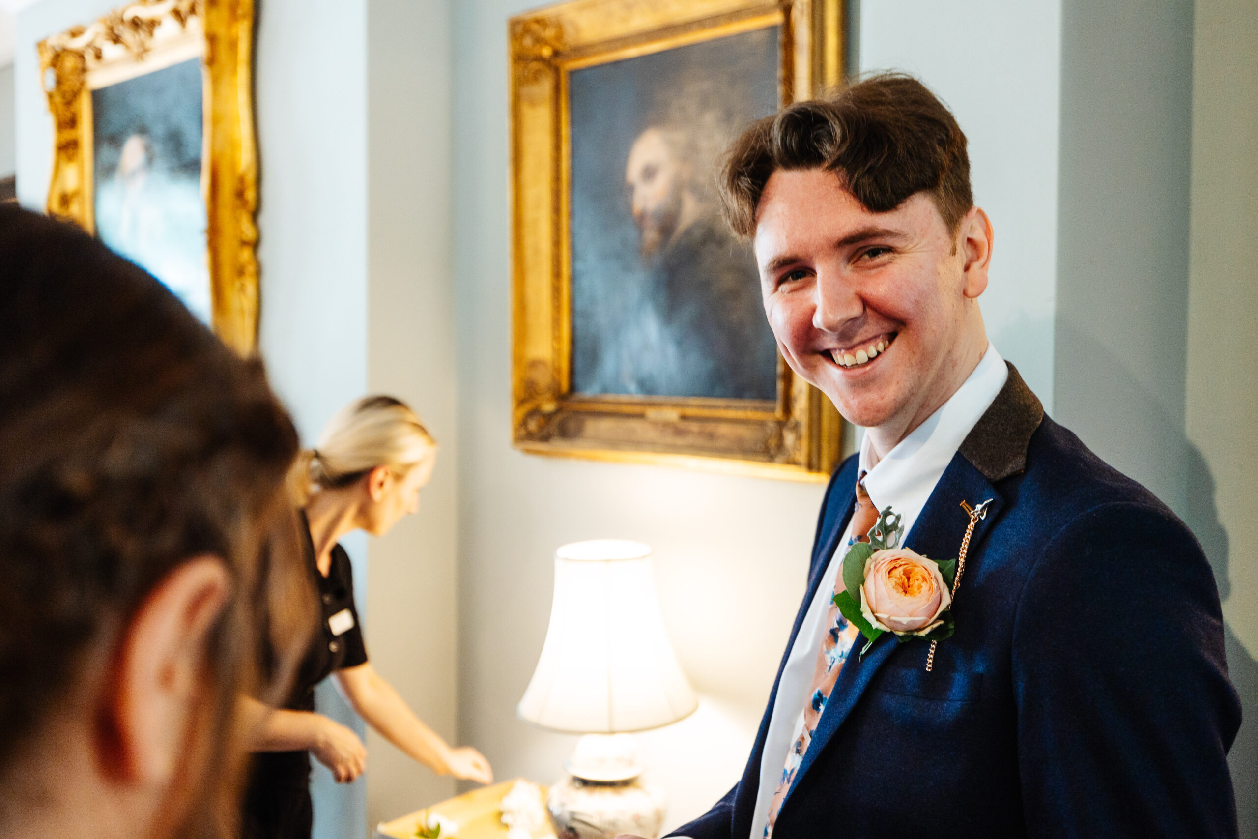 The groom, looking and smiling at the camera. He is in a navy suit and has a pale pink peony as his buttonhole.