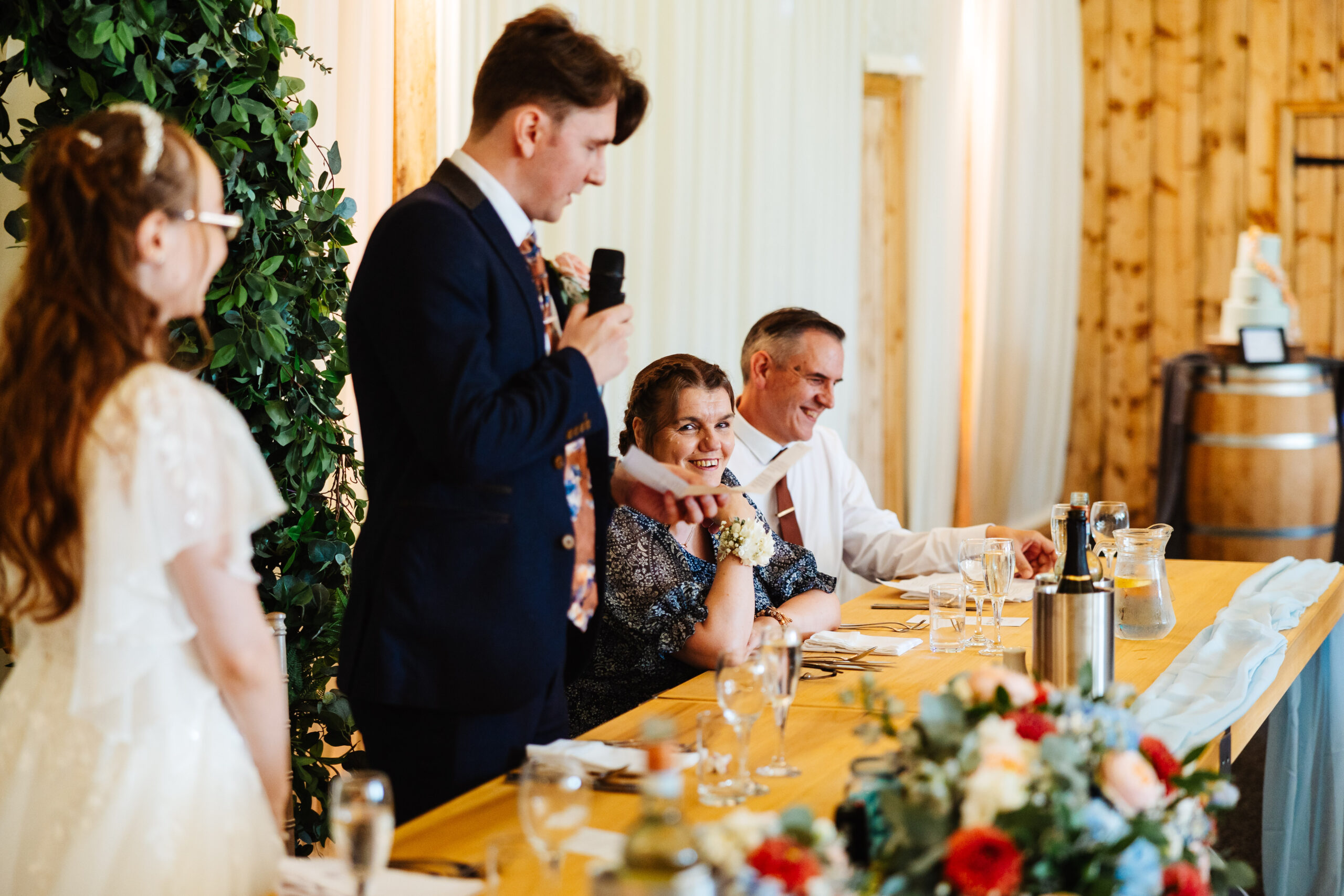 The groom is holding a microphone and looking at the guests. The bride is standing up during his speech.