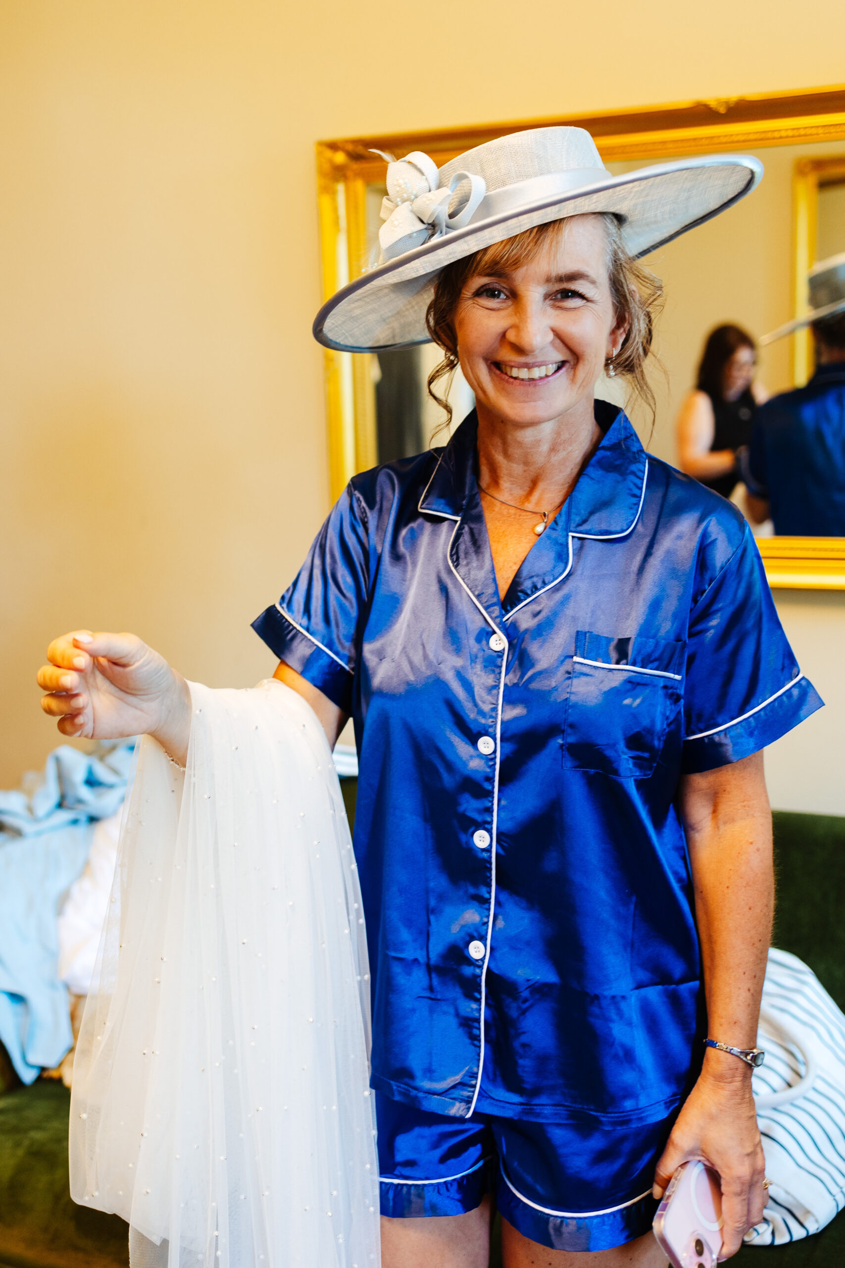 The mother of the bride. She is wearing navy pyjamas and a wide rimmed grey hat with a grey ribbon and fancy detailing on top. She is smiling.